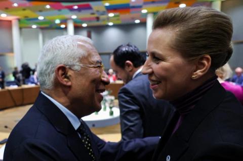 To personer i formelle antrekk smiler og snakker sammen ved et konferansebord i et sterkt opplyst møterom.