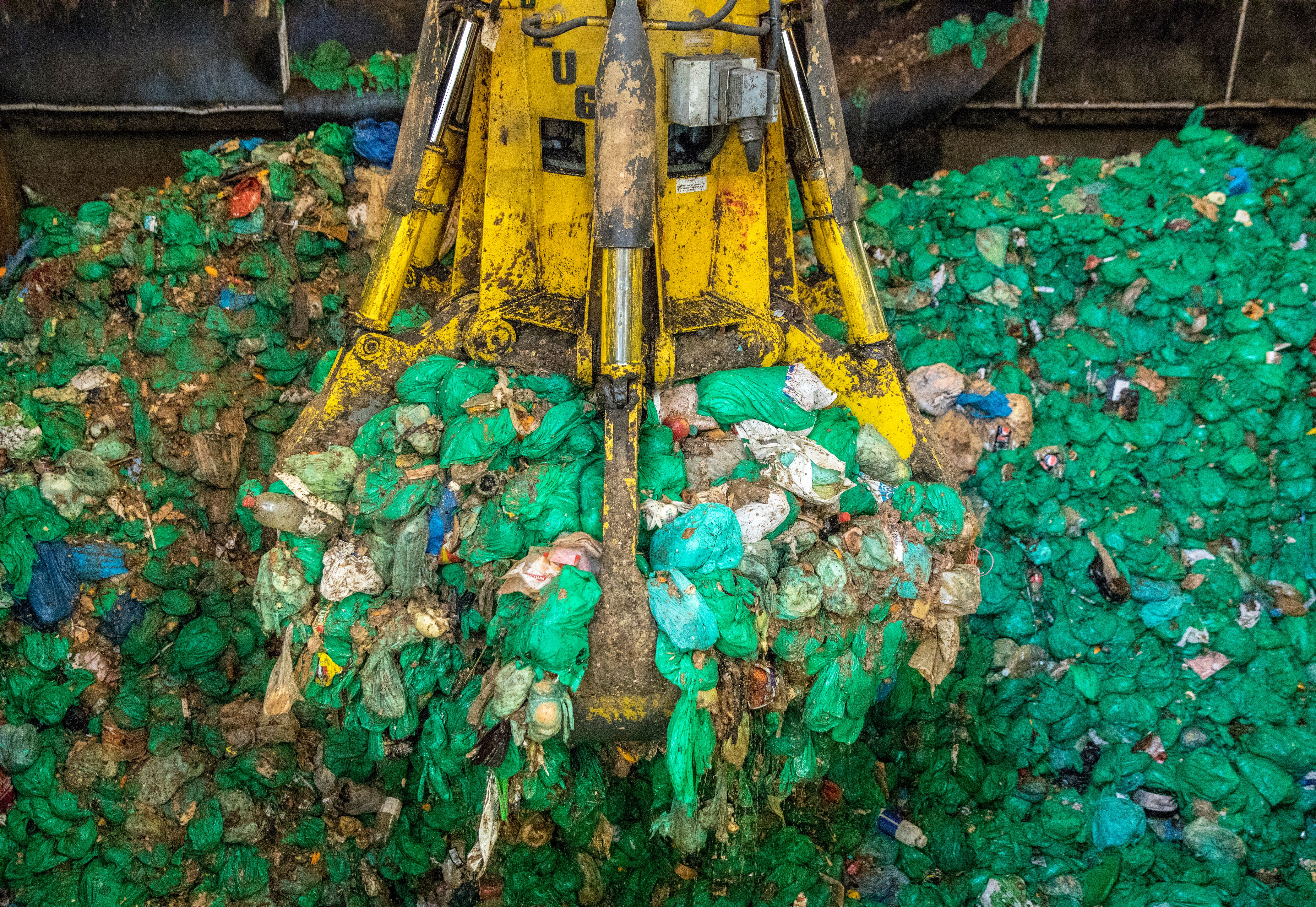 Industrial grabber moving a large pile of green plastic bags and mixed refuse.