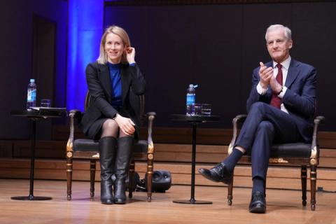 En kvinne i svart blazer og støvler sitter ved siden av en mann i dress på en scene, mannen klapper.