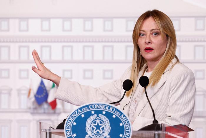 Kvinne i hvit blazer som taler fra en talerstol med mikrofoner, med venstre hånd hevet og håndflaten vendt ut.