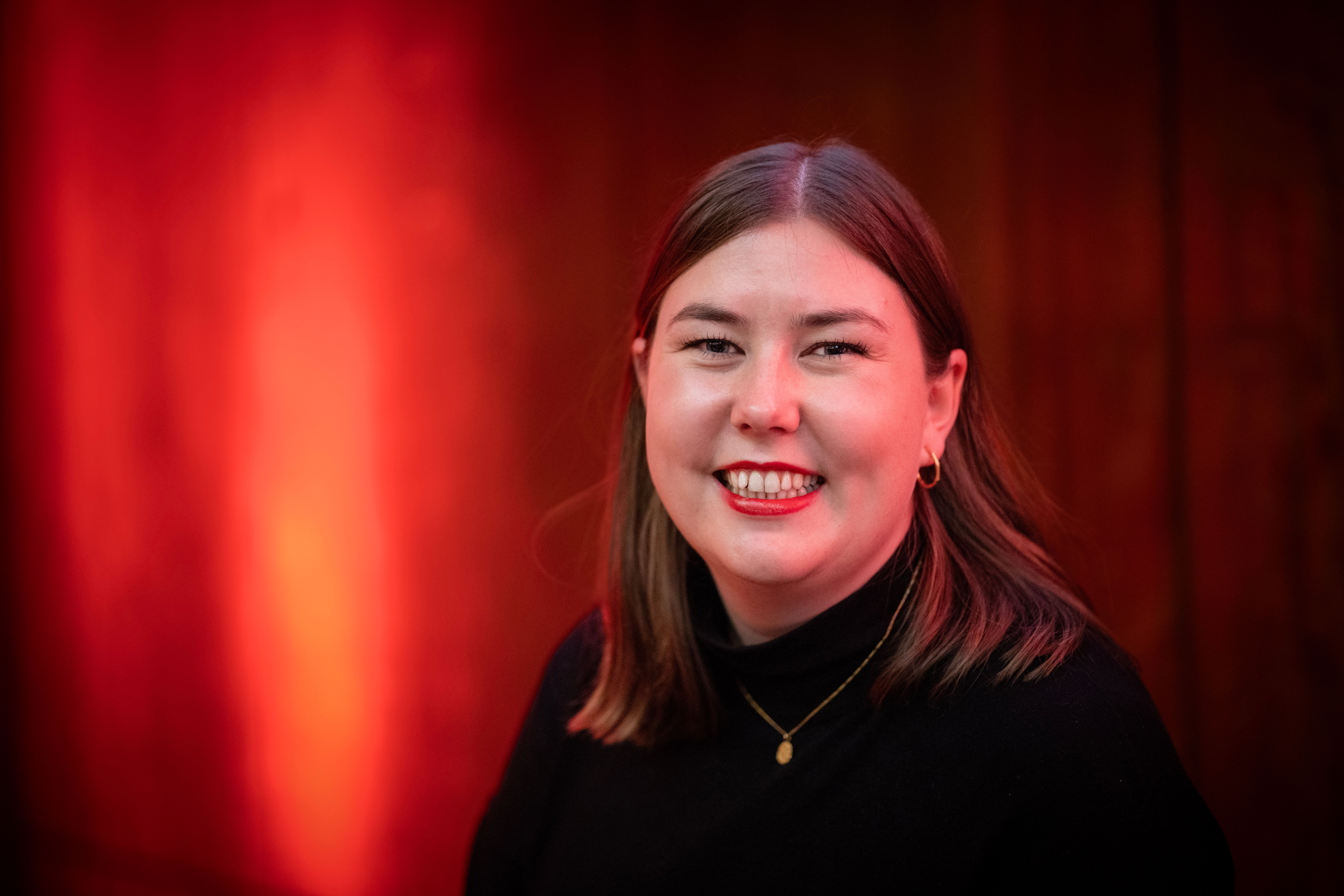 En smilende kvinne med brunt hår, iført en svart høyhalset genser og et gullkjede, står mot en rødoransje bakgrunn.