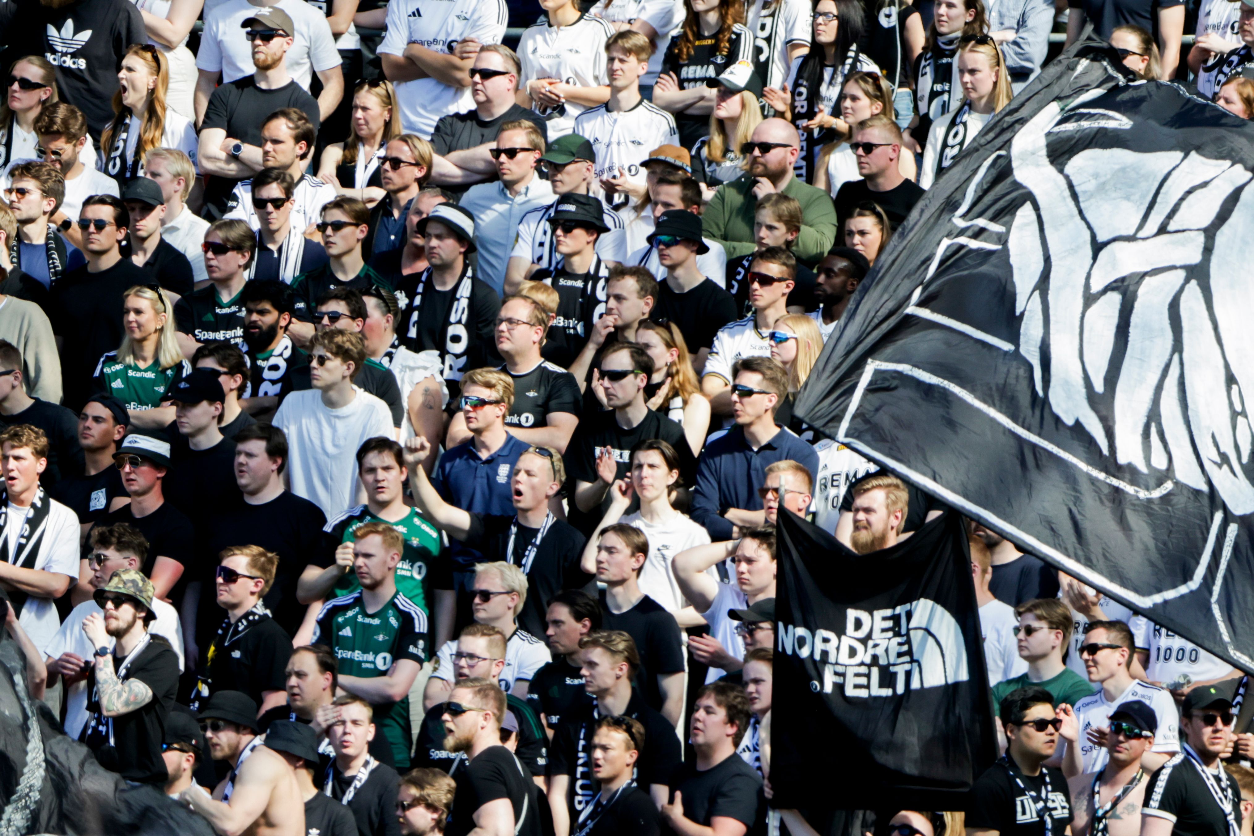 En stor folkemengde med sportsfans på tribunene på et stadion, noen holder svarte flagg med hvite motiver.