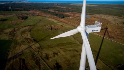 Fugleperspektiv av en Vestas-vindturbin som kaster en lang skygge over grønne jorder og skog, med havet i det fjerne.