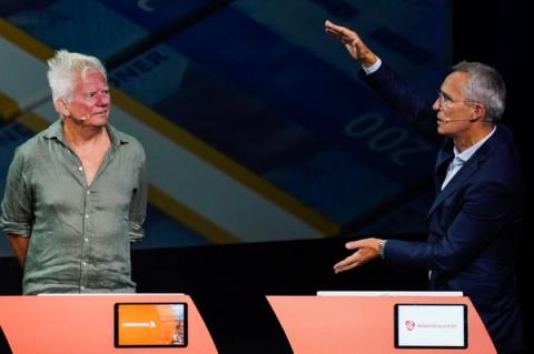 To menn på en debattscene, den ene i en uformell skjorte som ser på den andre i dress, som gestikulerer med hevede hender.