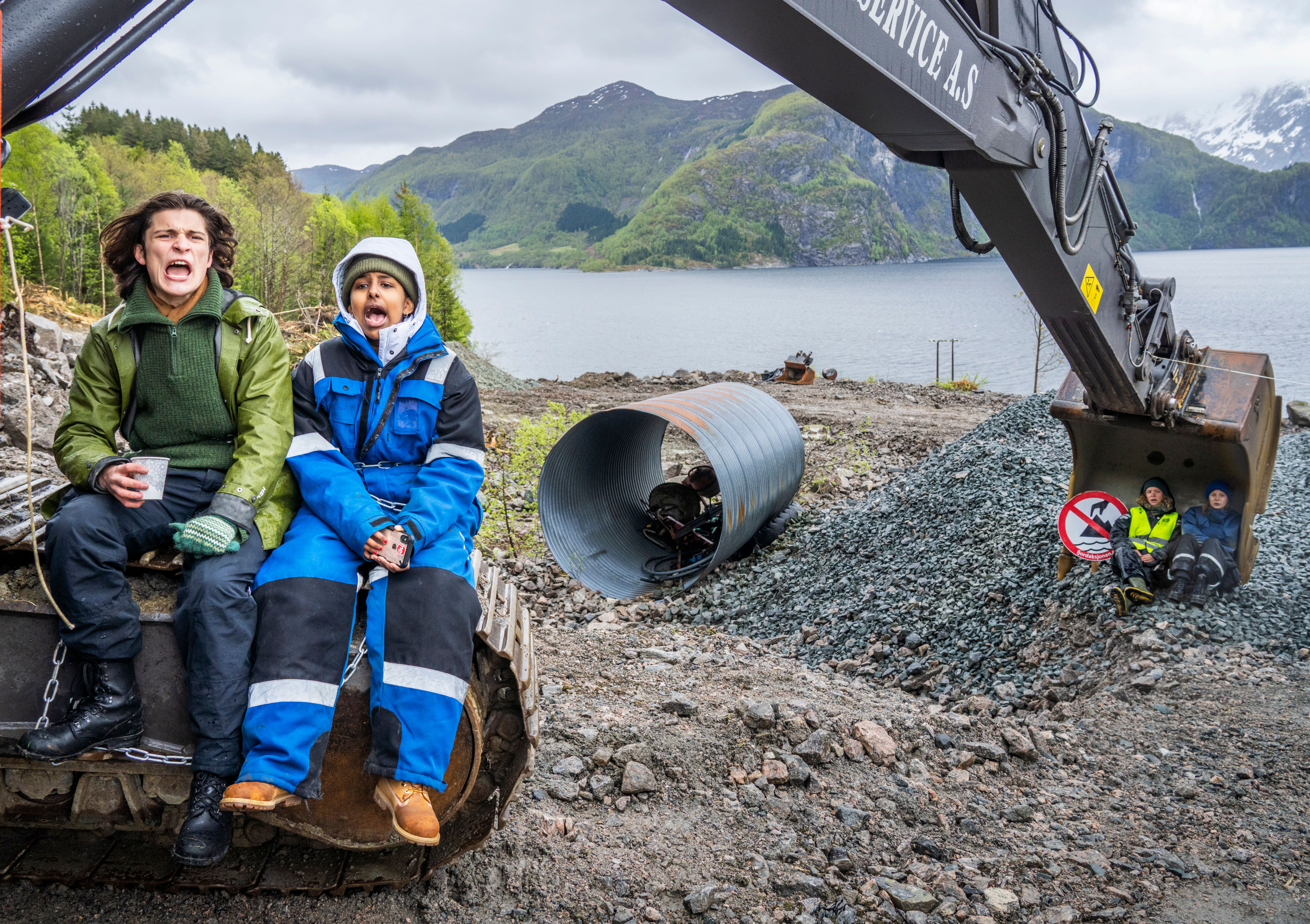 Two young people with wide open mouths sit on an excavator's tracks. Two others are in the excavator's bucket at a construction site by a body of water and mountains.