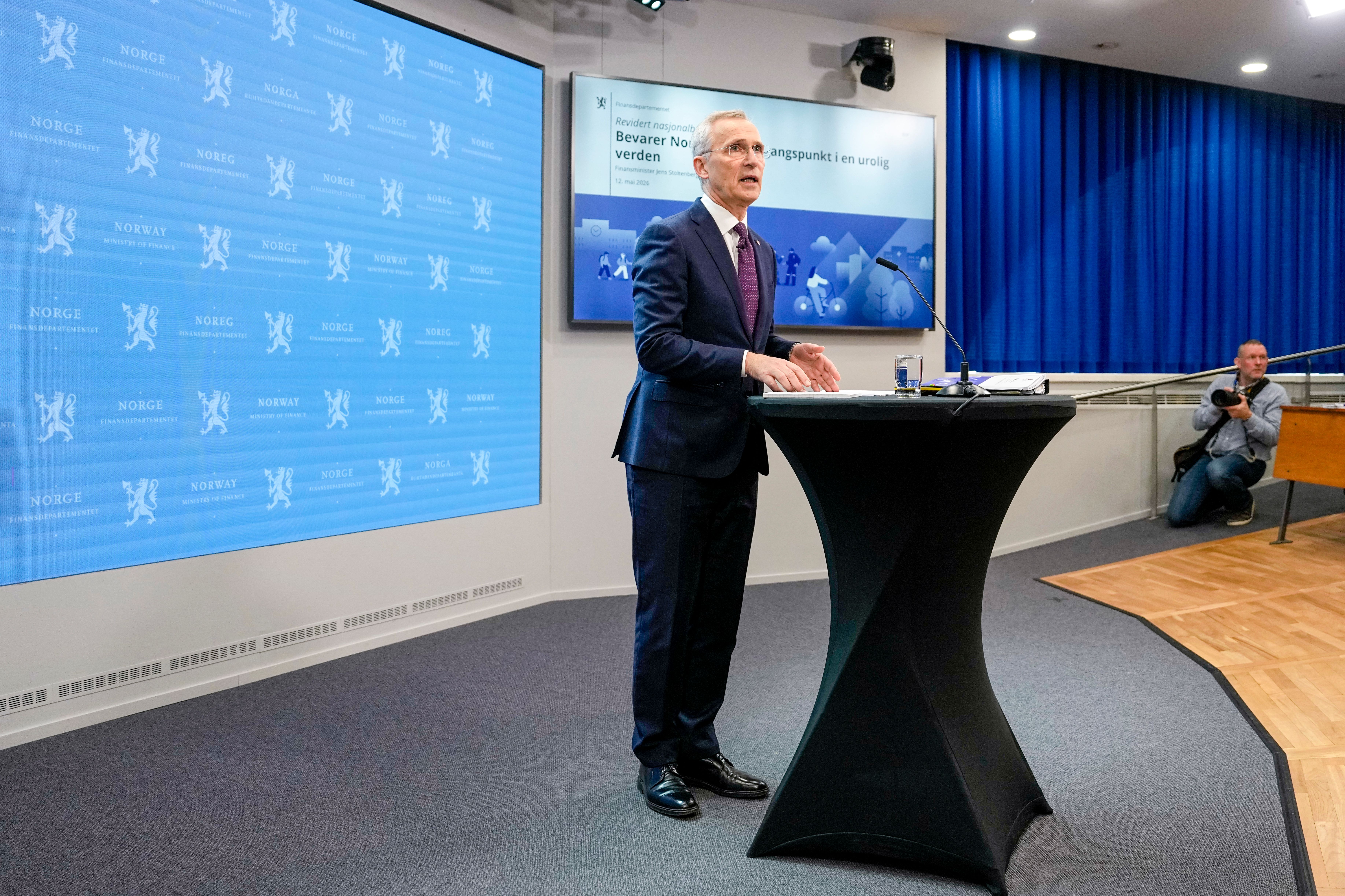 Mann i dress som taler fra en talerstol, flankert av skjermer som viser en presentasjon og norsk profilering.