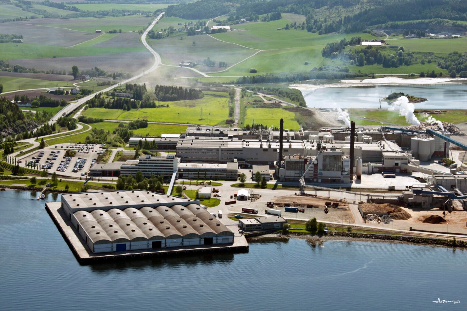 Flyfoto av et stort industrikompleks med skorsteiner og en kai ved en elv, omgitt av grønt landskap.