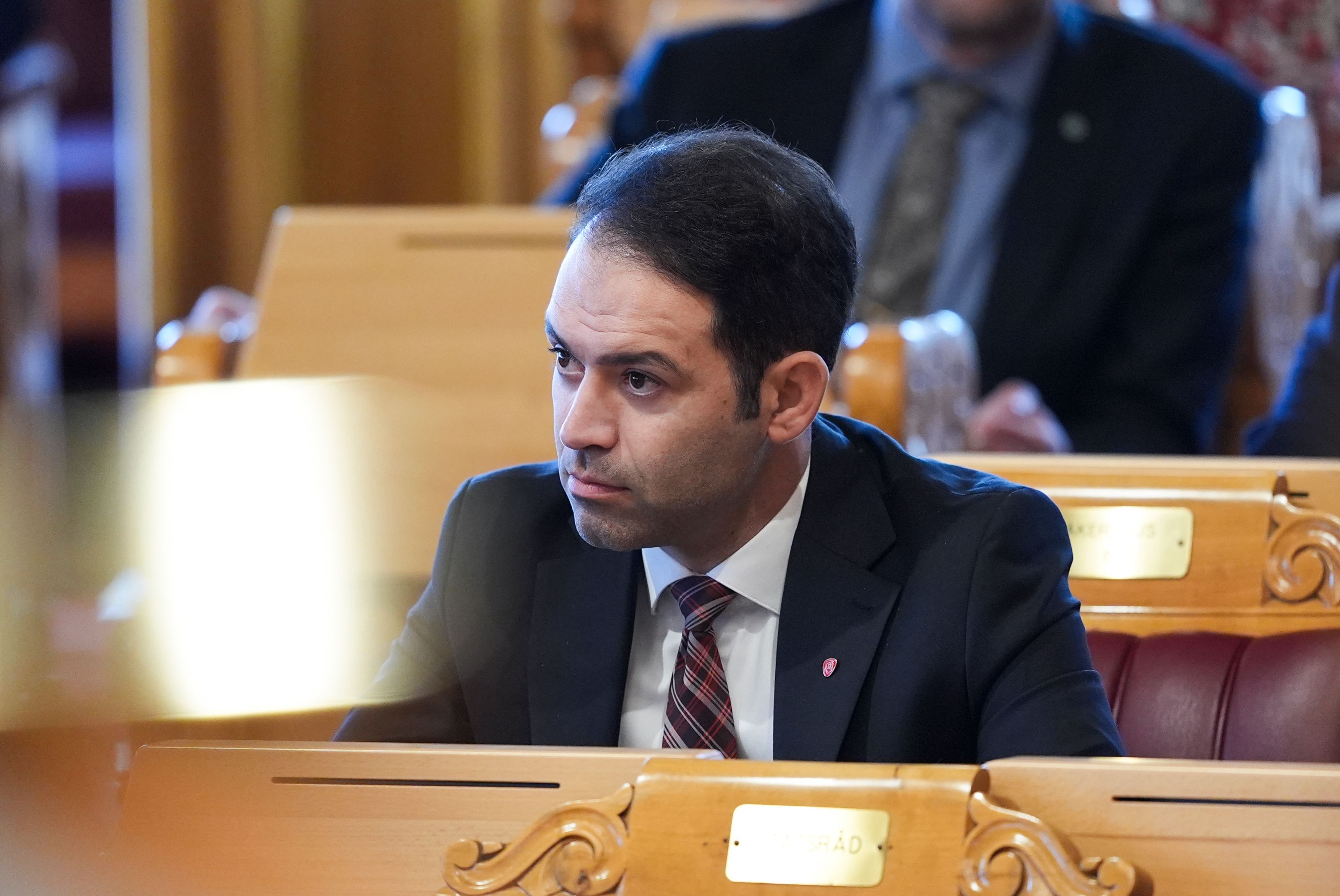 En mann i dress og slips sitter ved et trebord i en formell setting og ser intenst til siden.