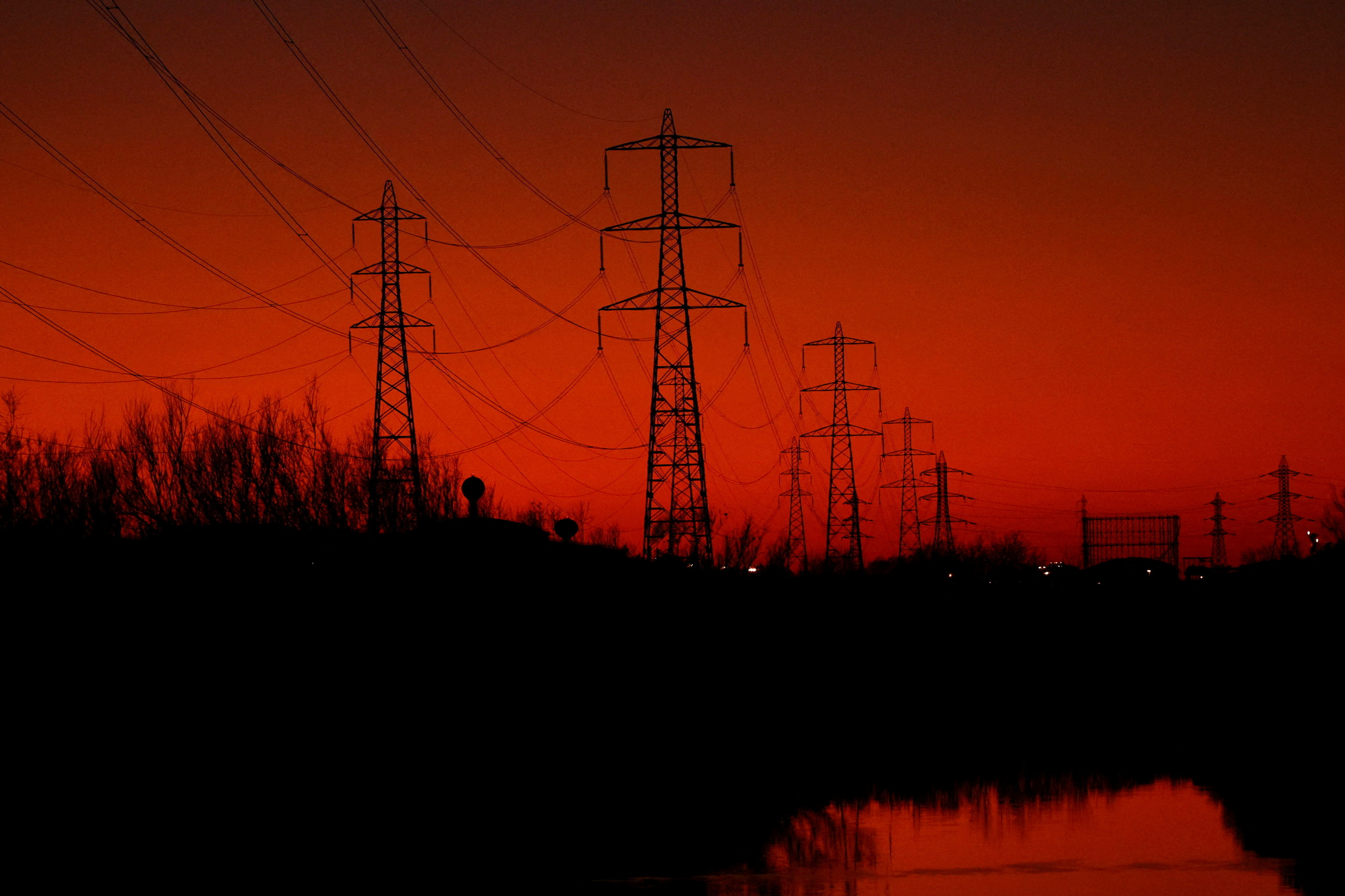 Silhuettaktige kraftlinjetårn og trær speilet i vannet under en dramatisk rød himmel.