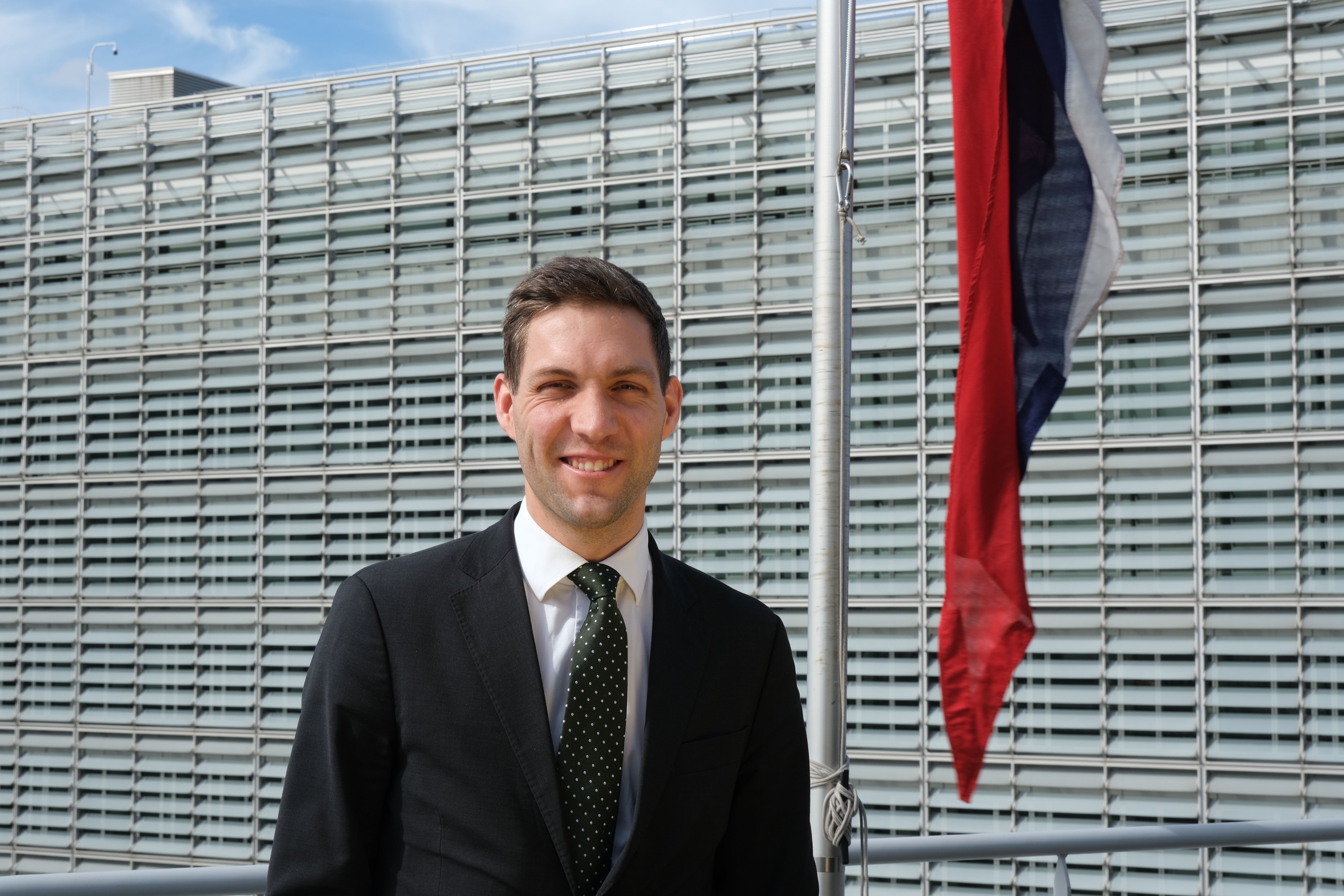 Smiling man in a suit stands outside a modern building with a flag.