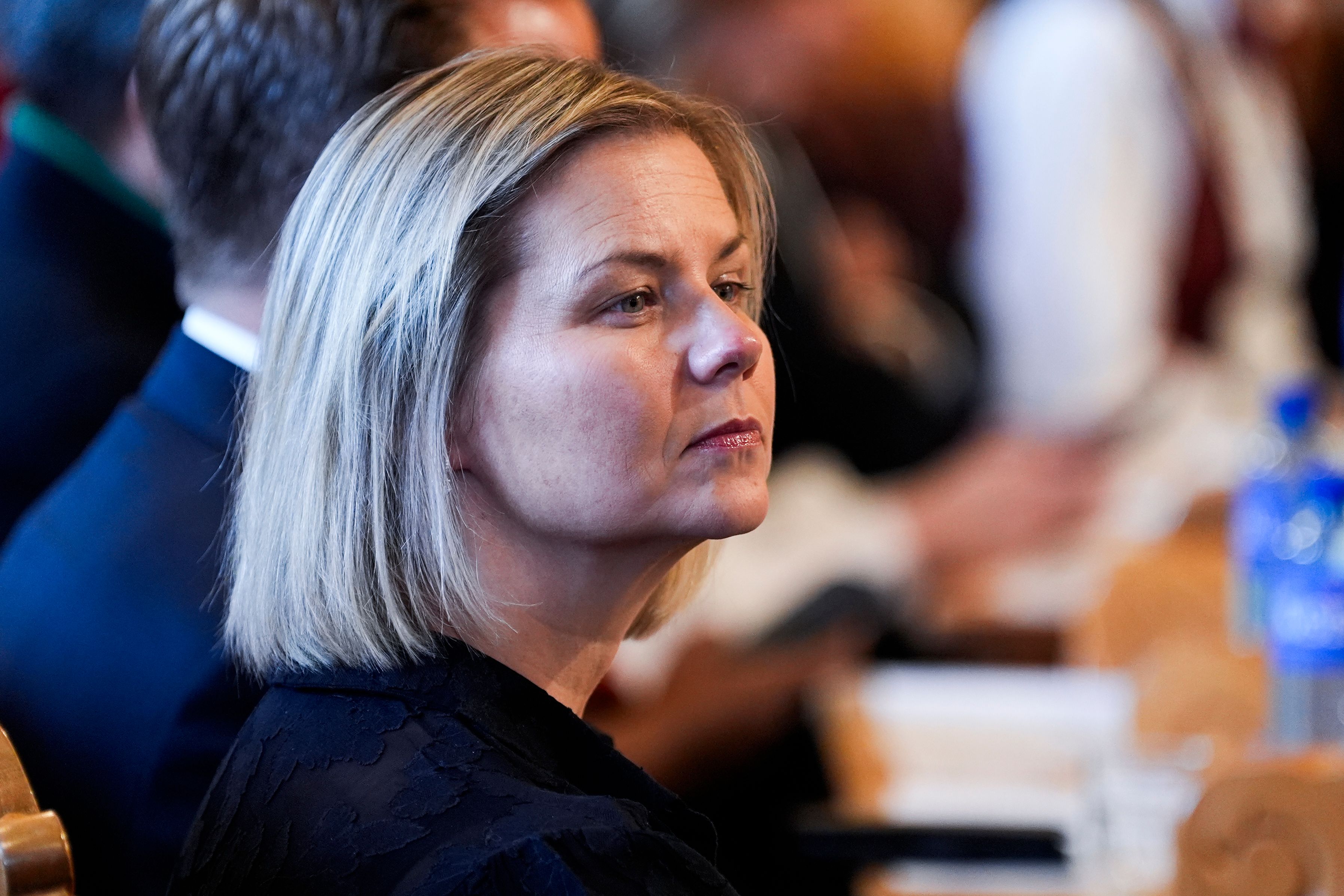 En blond kvinne med alvorlig uttrykk ser mot høyre, med en uklar bakgrunn av mennesker.