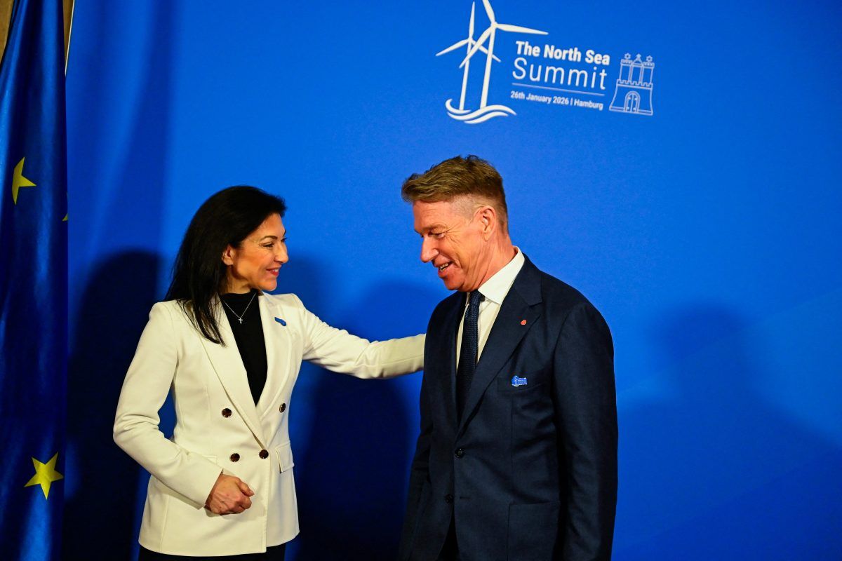 To personer i formelle antrekk hilser på hverandre foran et blått bakteppe merket "The North Sea Summit" med detaljer og grafikk om arrangementet. Et EU-flagg er delvis synlig til venstre.