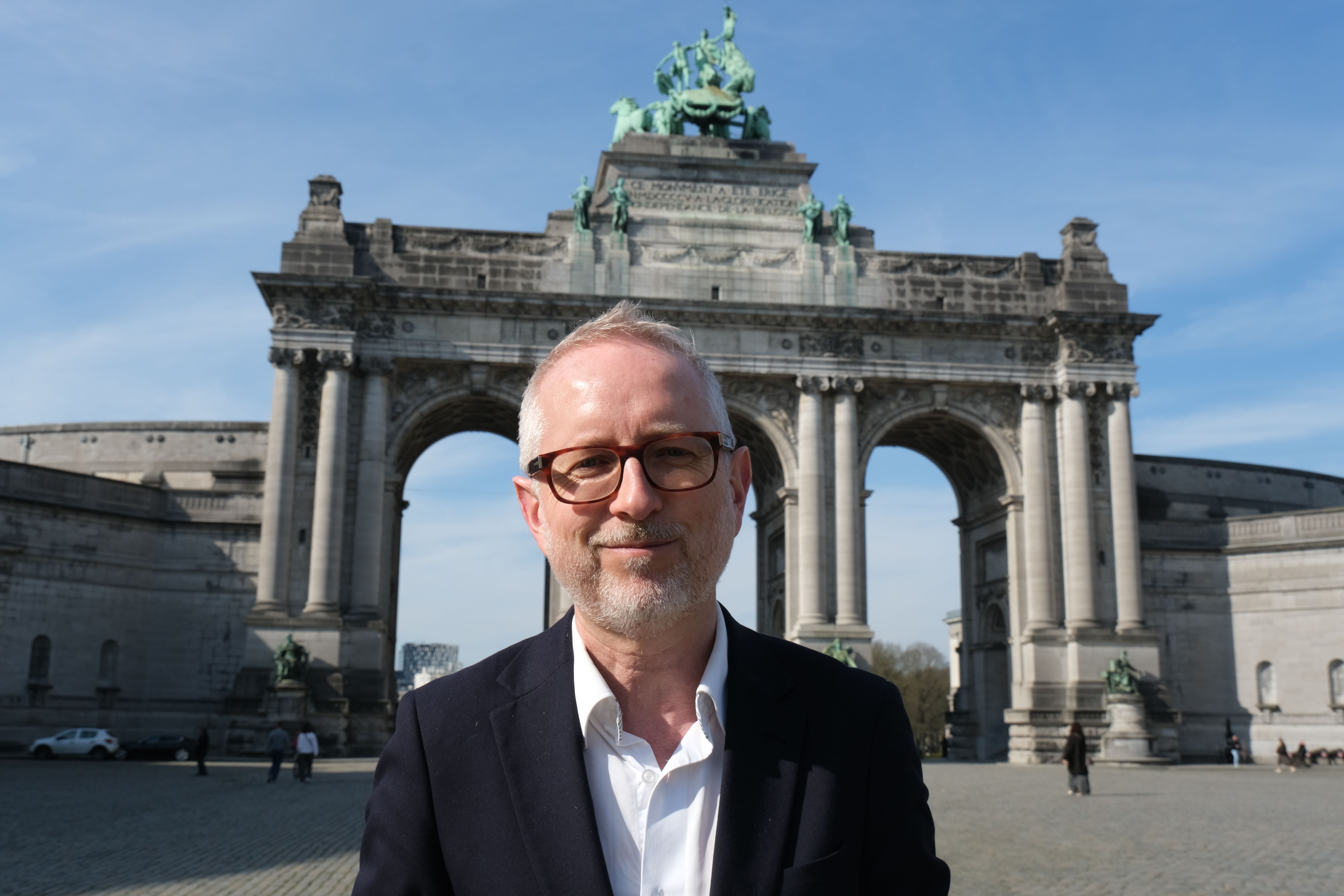 En mann med briller og skjegg smiler foran Cinquantenaire-buen i Brussel.