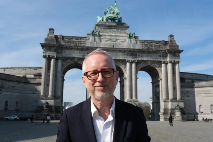 En mann med briller og skjegg smiler foran Cinquantenaire-buen i Brussel.