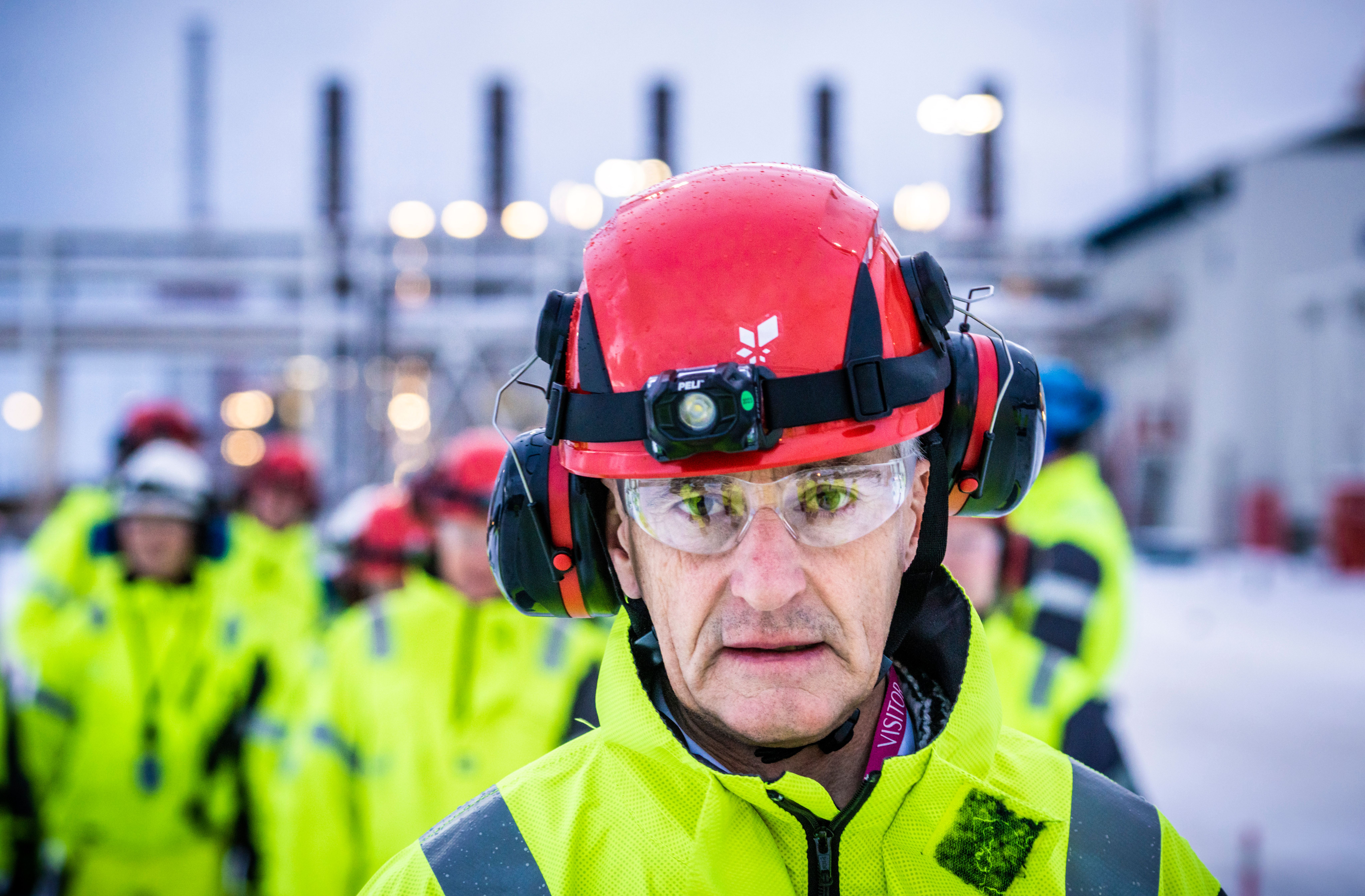 Mann med rød vernehjelm, vernebriller og gul høytsynlig jakke på et snødekt industriområde.