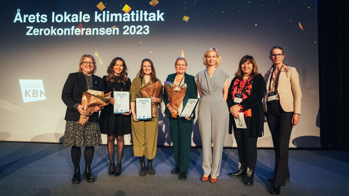 Syv personer står smilende på en scene med blomsterbuketter og diplomer i hendene