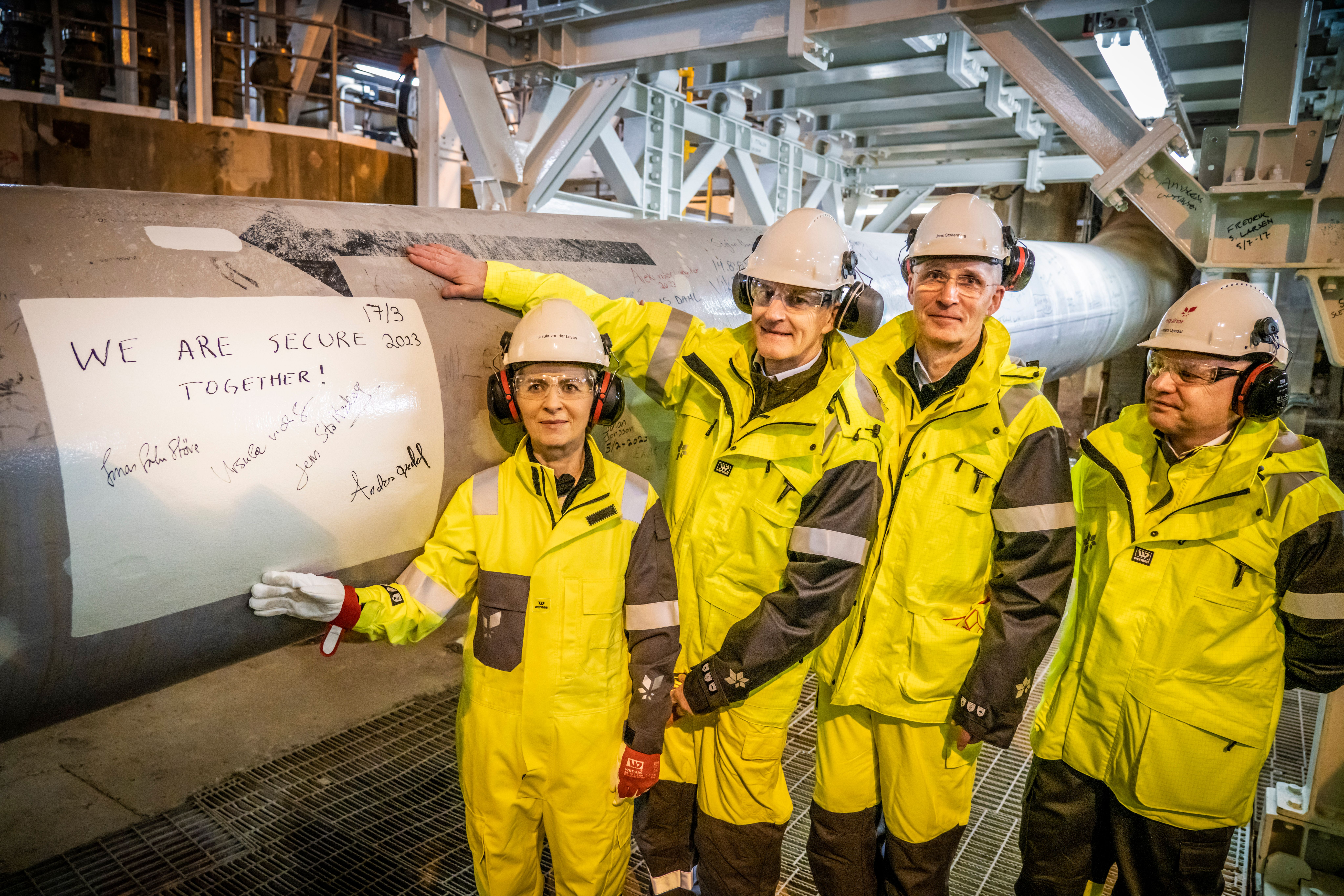 Fire personer i verneutstyr ved siden av et stort industrirør, der et ark med teksten «VI ER TRYGGERE SAMMEN!» med signaturer og datoen 2017 er festet.