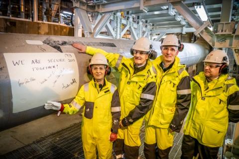 Fire personer i verneutstyr ved siden av et stort industrirør, der et ark med teksten «VI ER TRYGGERE SAMMEN!» med signaturer og datoen 2017 er festet.