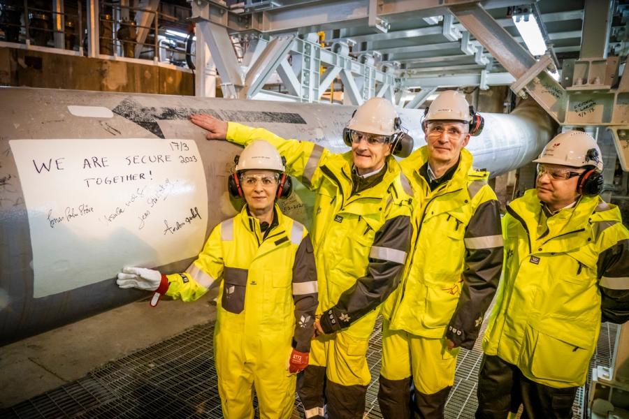 Fire personer i verneutstyr ved siden av et stort industrirør, der et ark med teksten «VI ER TRYGGERE SAMMEN!» med signaturer og datoen 2017 er festet.