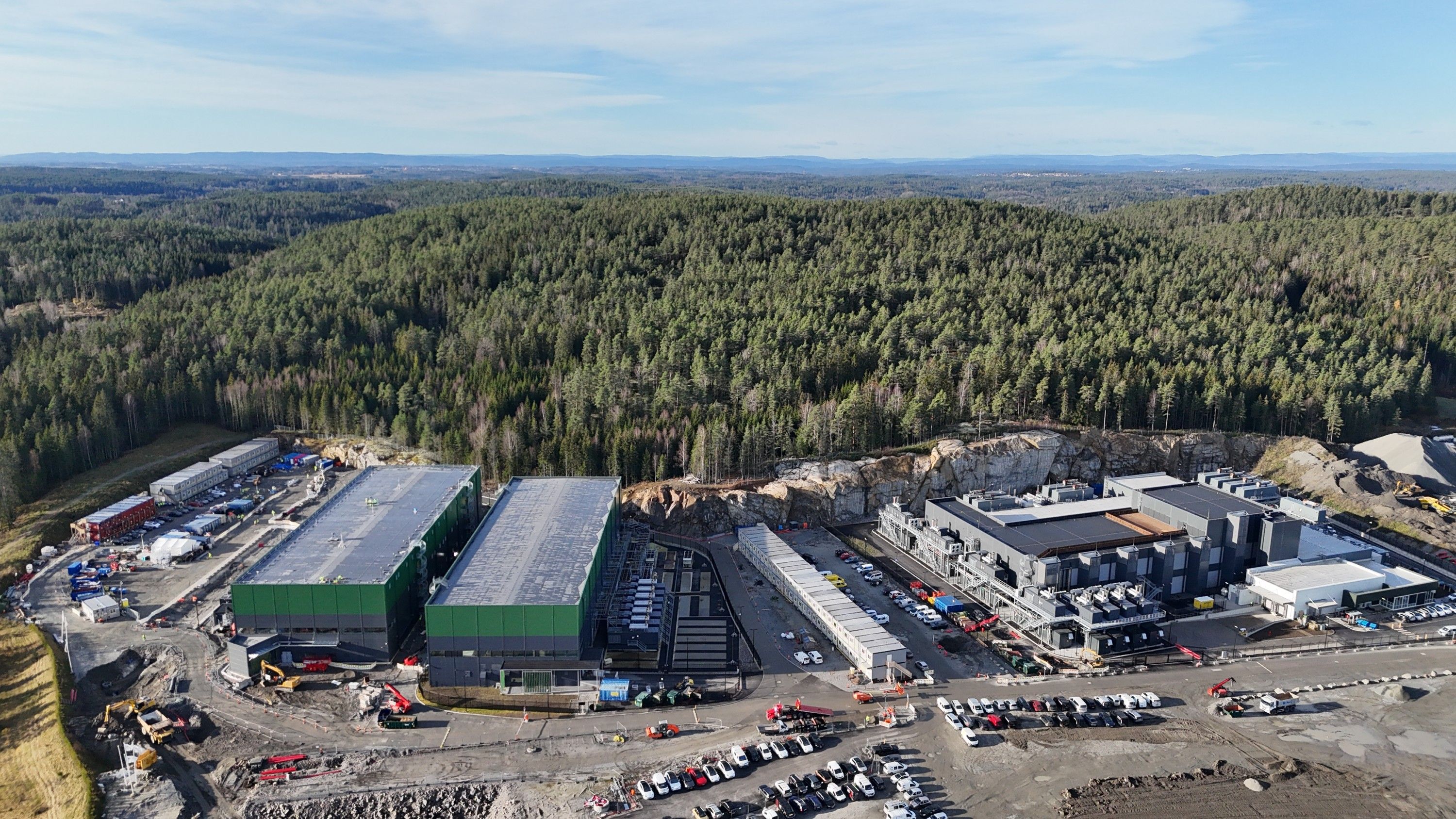 Flyfoto av et stort datasenterkompleks med grønne og grå bygninger, omgitt av tett skog.