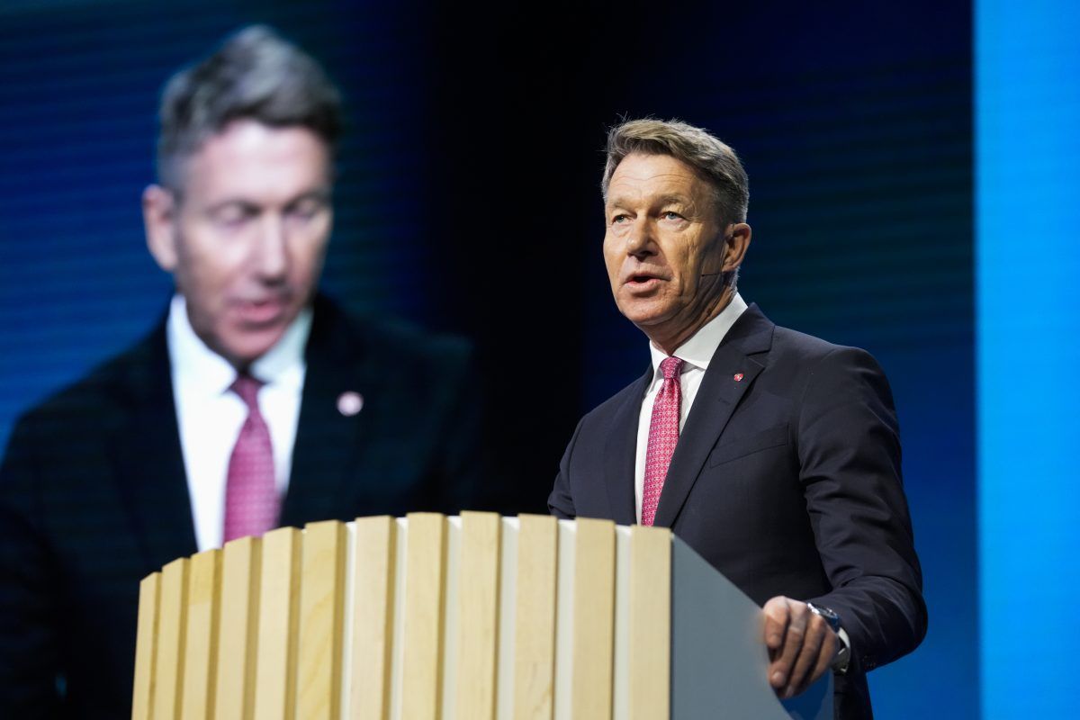 Mann i dress som taler fra en talerstol, med sitt uklare speilbilde på en skjerm bak seg.