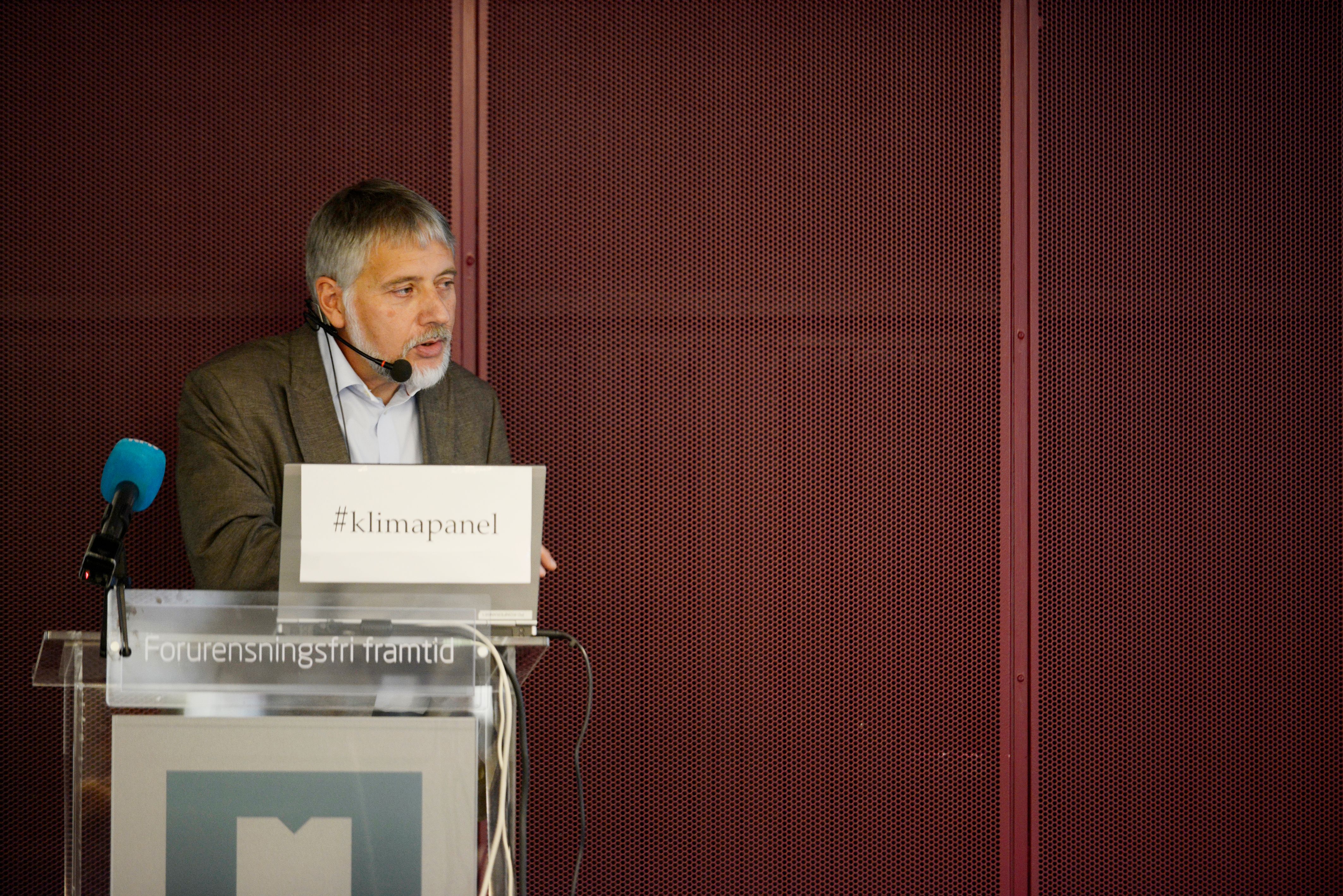 Professor Eystein Jansen taler fra en talerstol med mikrofoner og skilt med teksten «#klimapanel» og «Forurensningsfri framtid».