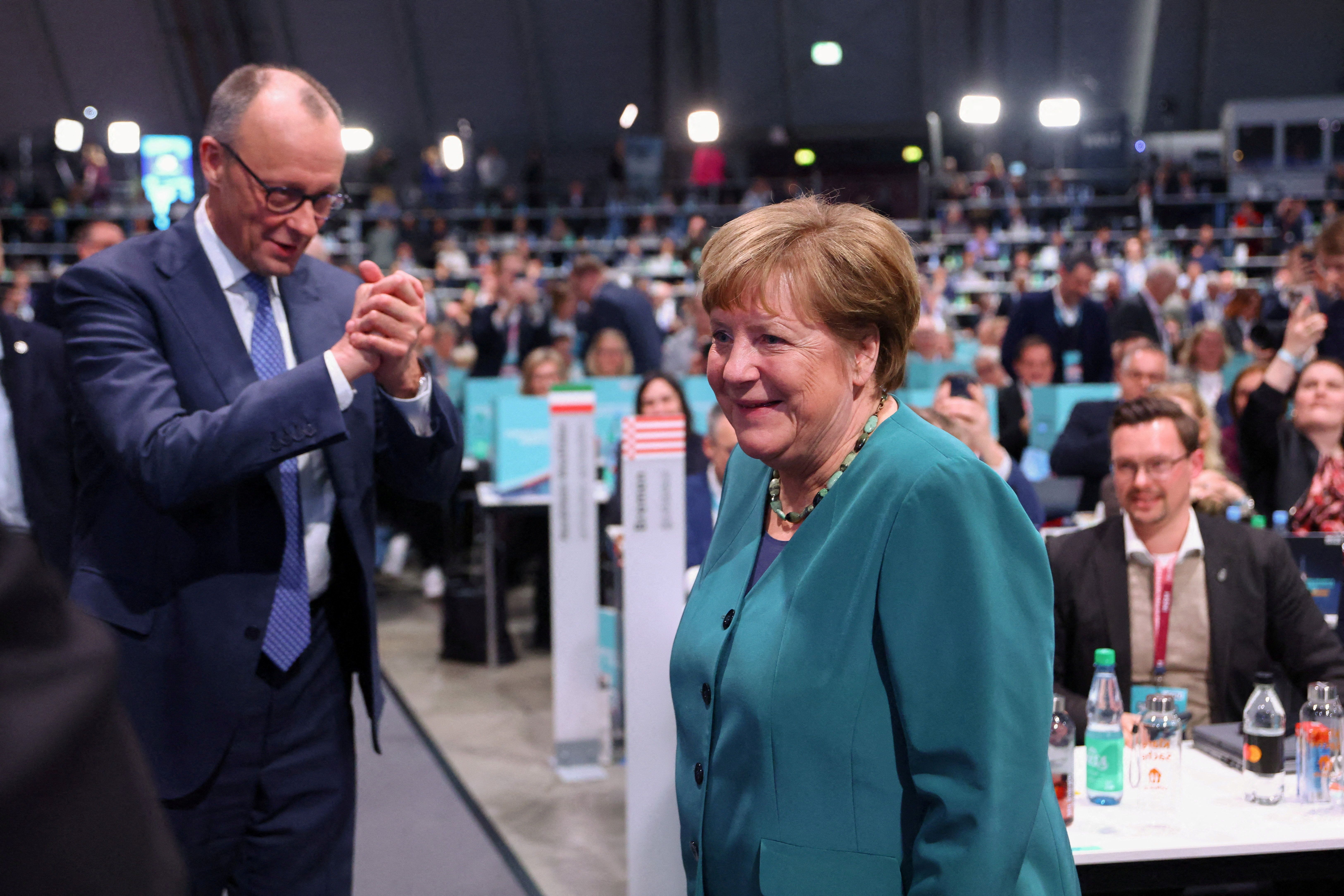 Angela Merkel smiler i en turkis jakke mens Friedrich Merz applauderer henne på et arrangement.