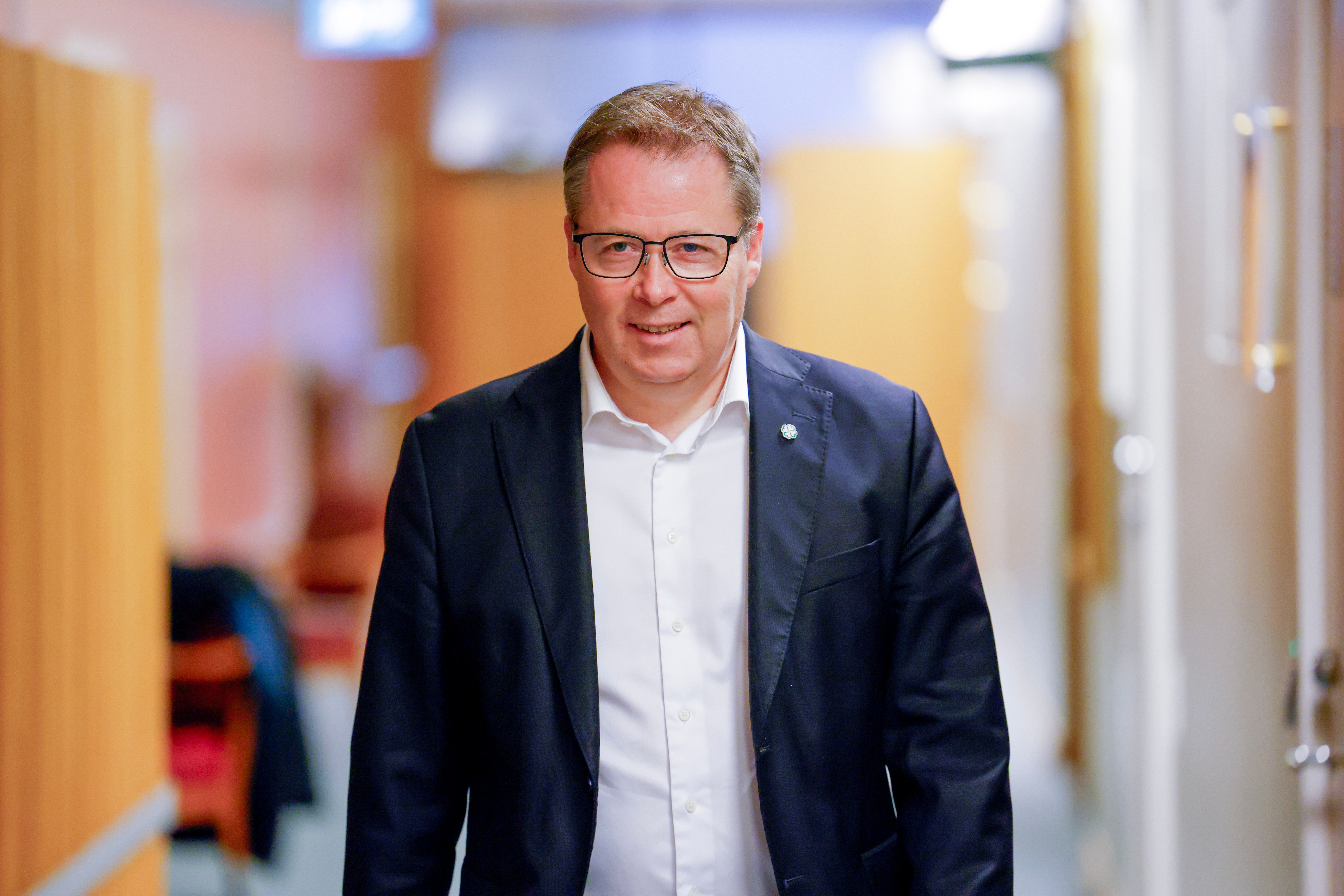 Bildet viser nestleder i Senterpartiet, Bjørn Arild Gram, i dress og briller som går bortover og smiler i en korridor.