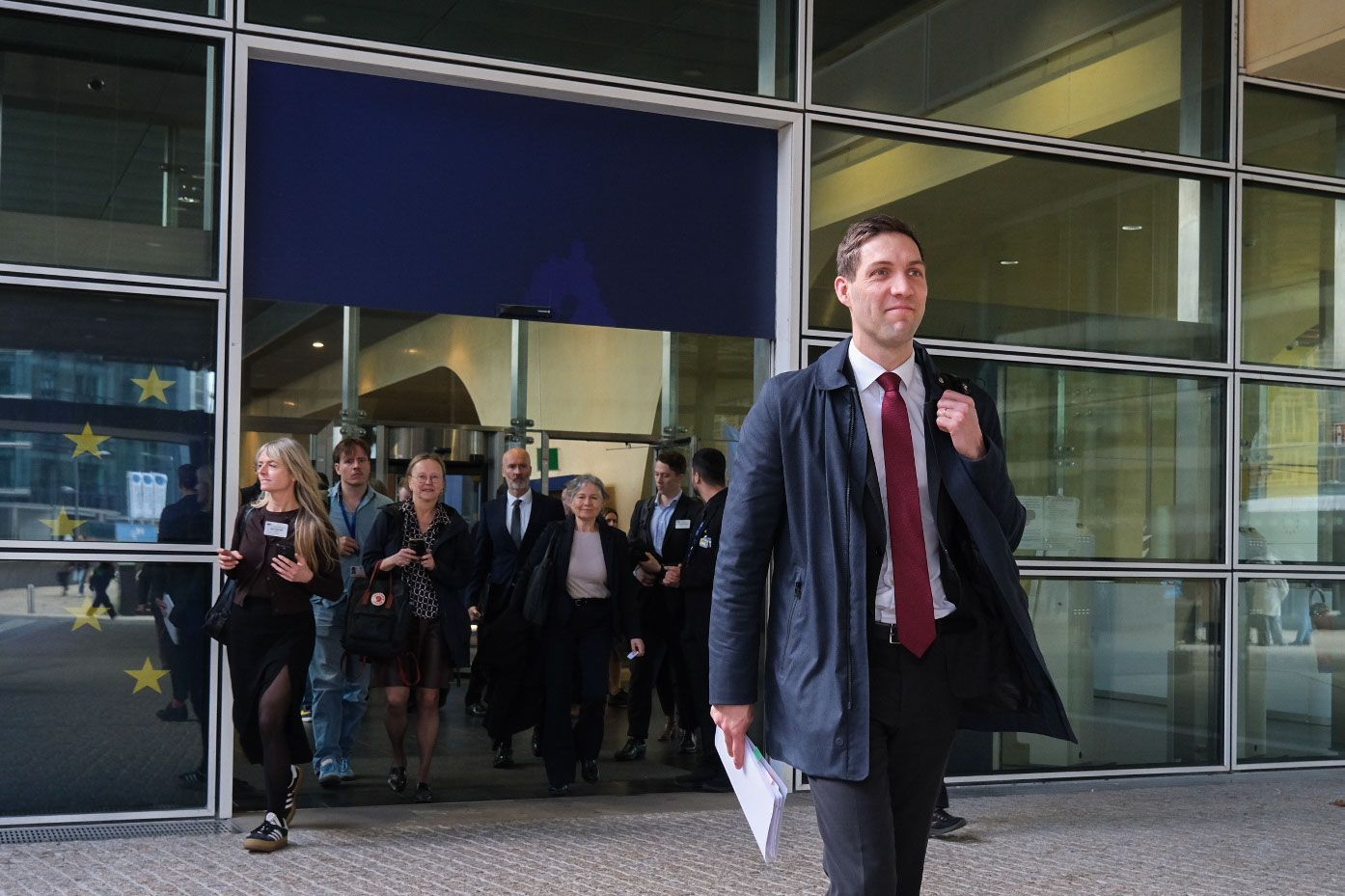 En mann i frakk og dress kommer ut av en moderne bygning med noen papirer i hånden, med andre personer bak seg.