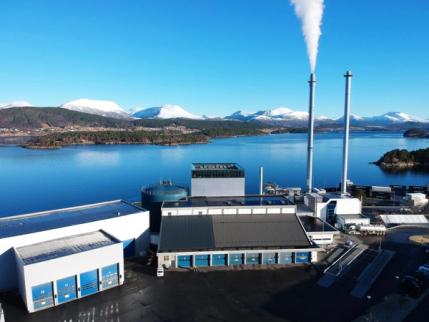 Industrianlegg ved vannkanten med to skorsteiner, hvorav den ene slipper ut røyk, med snødekte fjell i bakgrunnen.