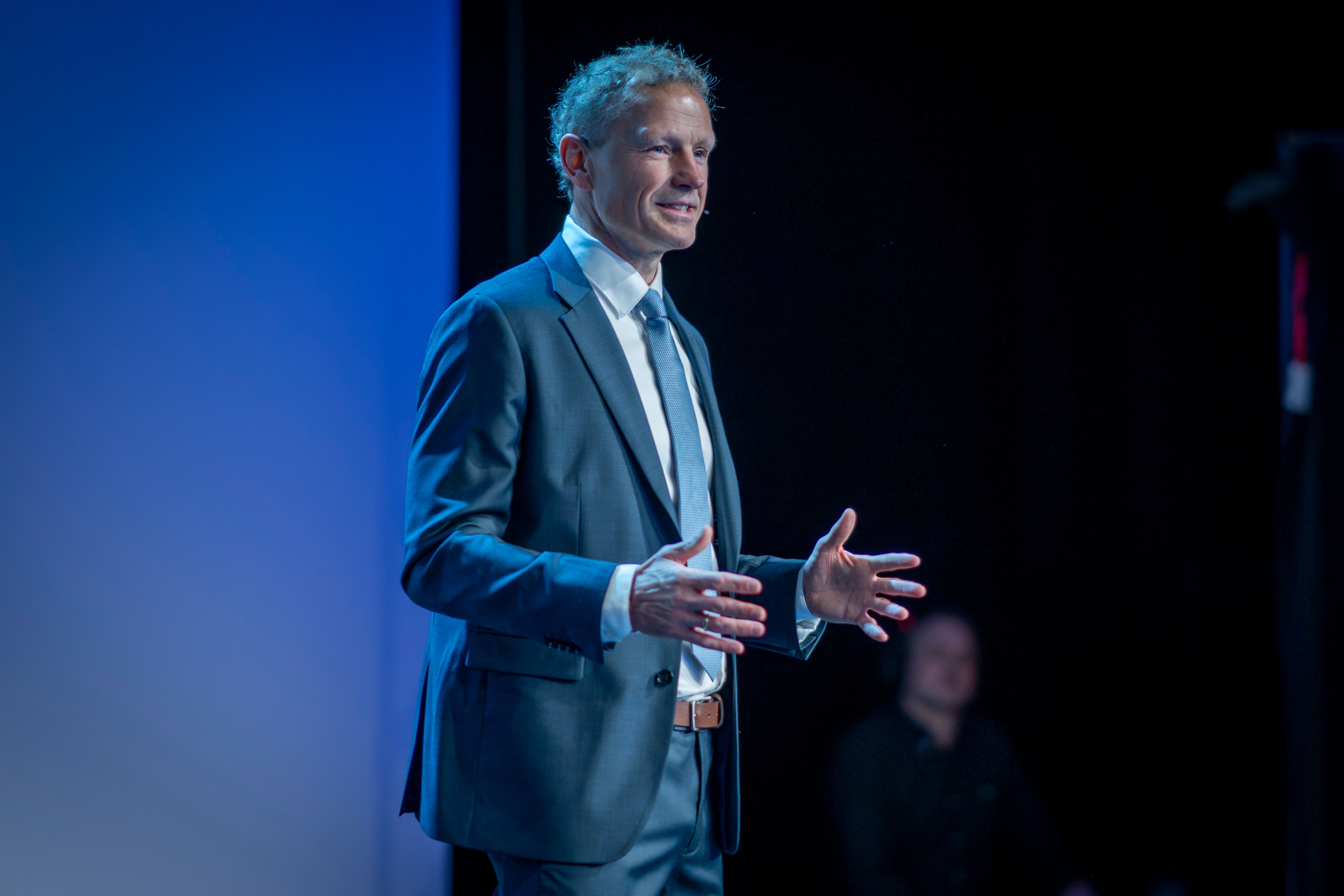 En mann i dress smiler og gestikulerer mens han presenterer på en scene med blå belysning.
