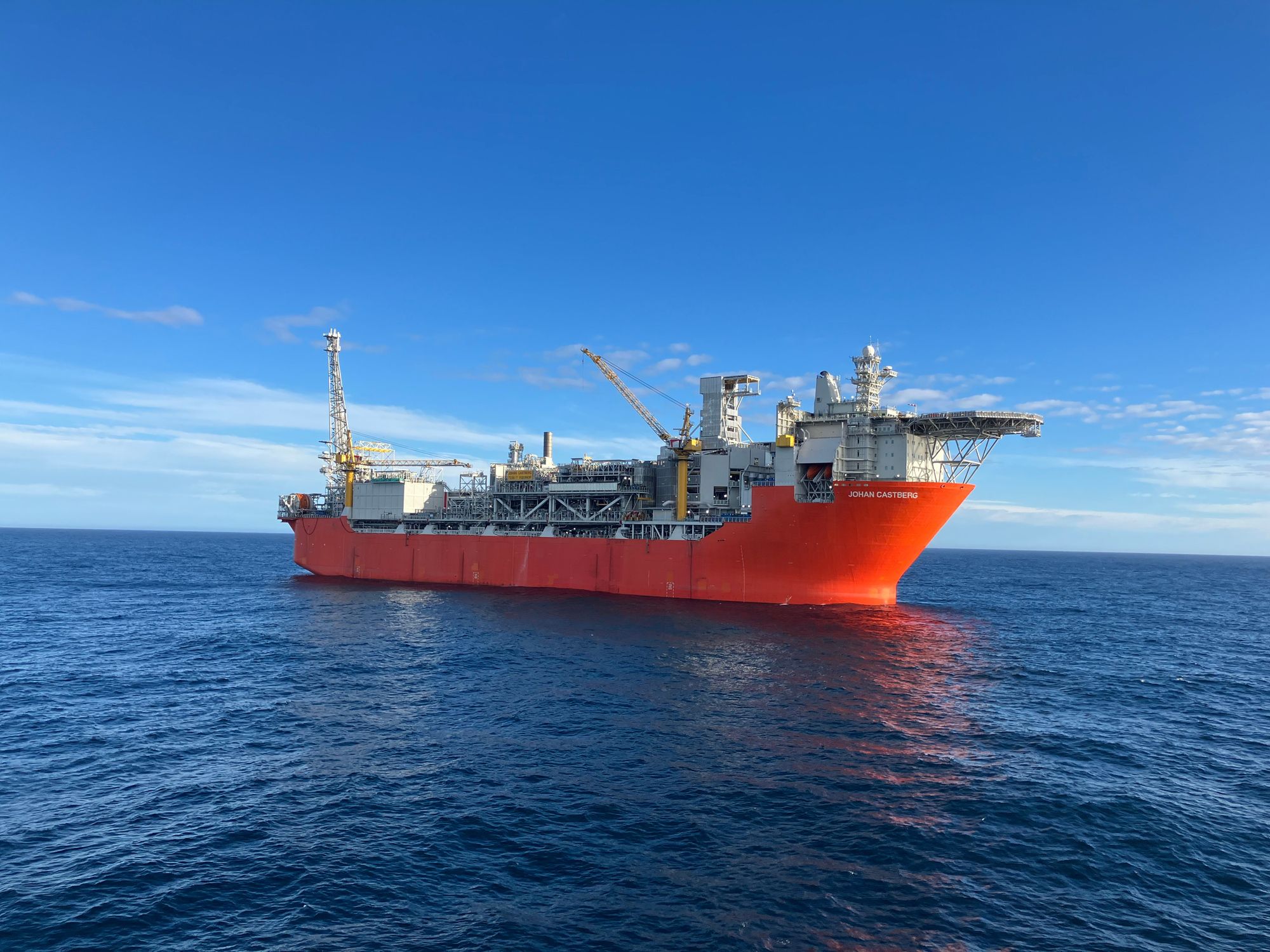 Et stort oransjerødt offshore produksjonsskip flyter på rolig blått vann under en klar blå himmel.