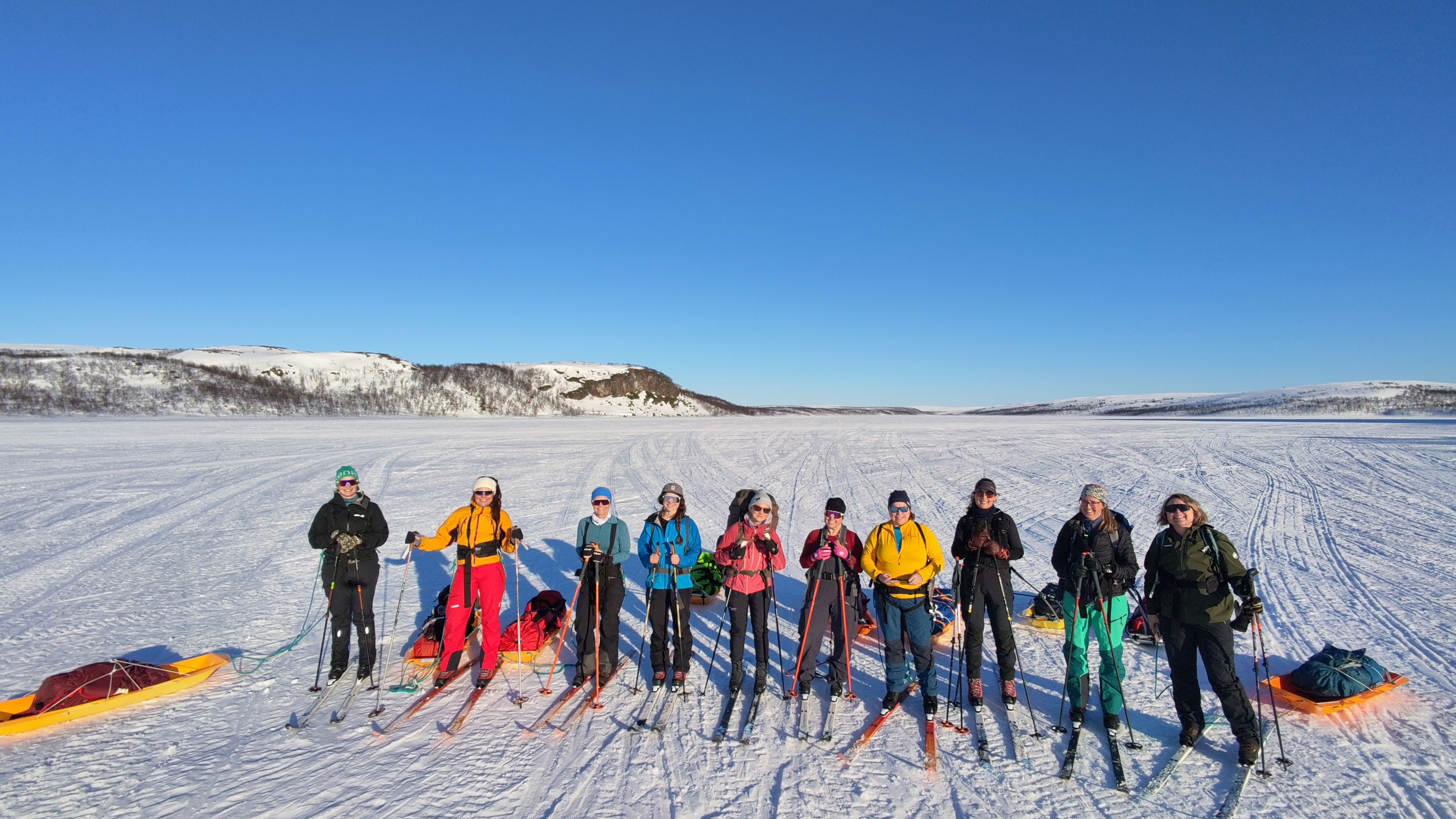 Ni personer på ski med pulker i et snødekt landskap under en klar blå himmel med åser i det fjerne.