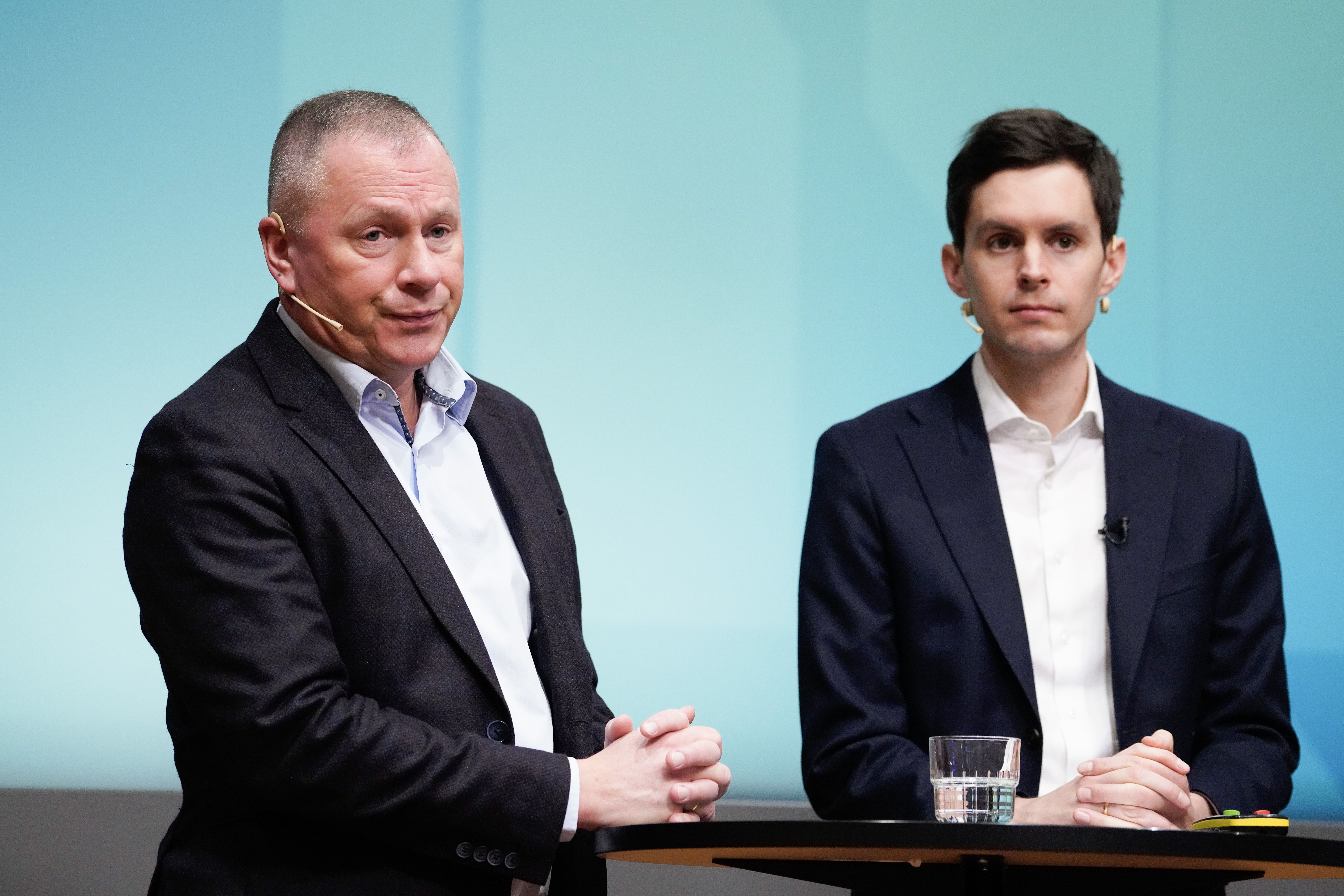 To menn i dress står ved et bord mot en lyseblå bakgrunn.