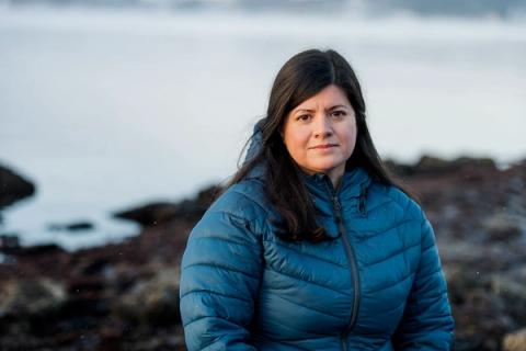 En kvinne i en blå boblejakke står ved en steinete strand med vann i bakgrunnen og ser mot kameraet.