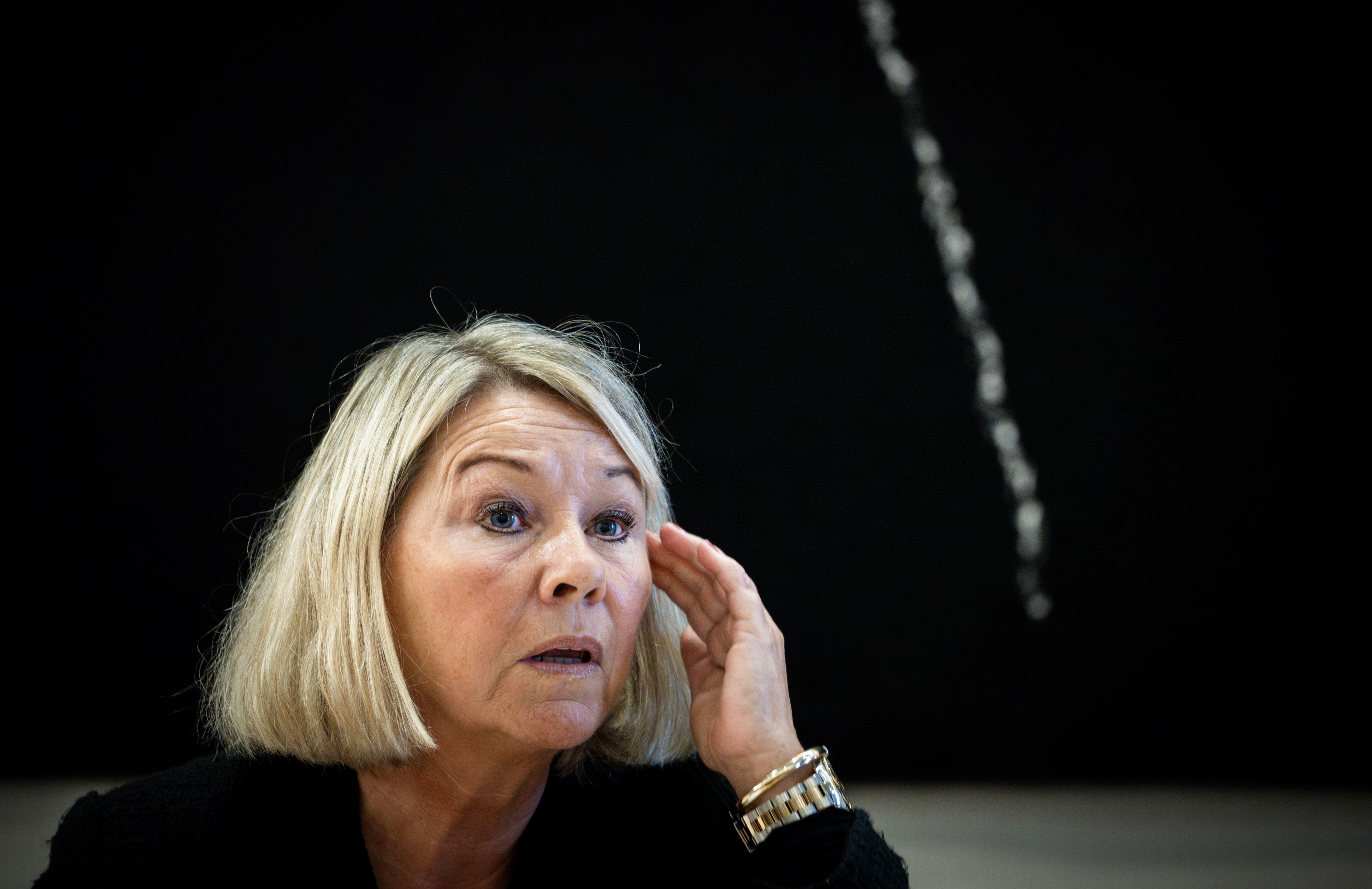 En blond kvinne med kort bobfrisyre ser overrasket ut og holder venstre hånd mot tinningen.