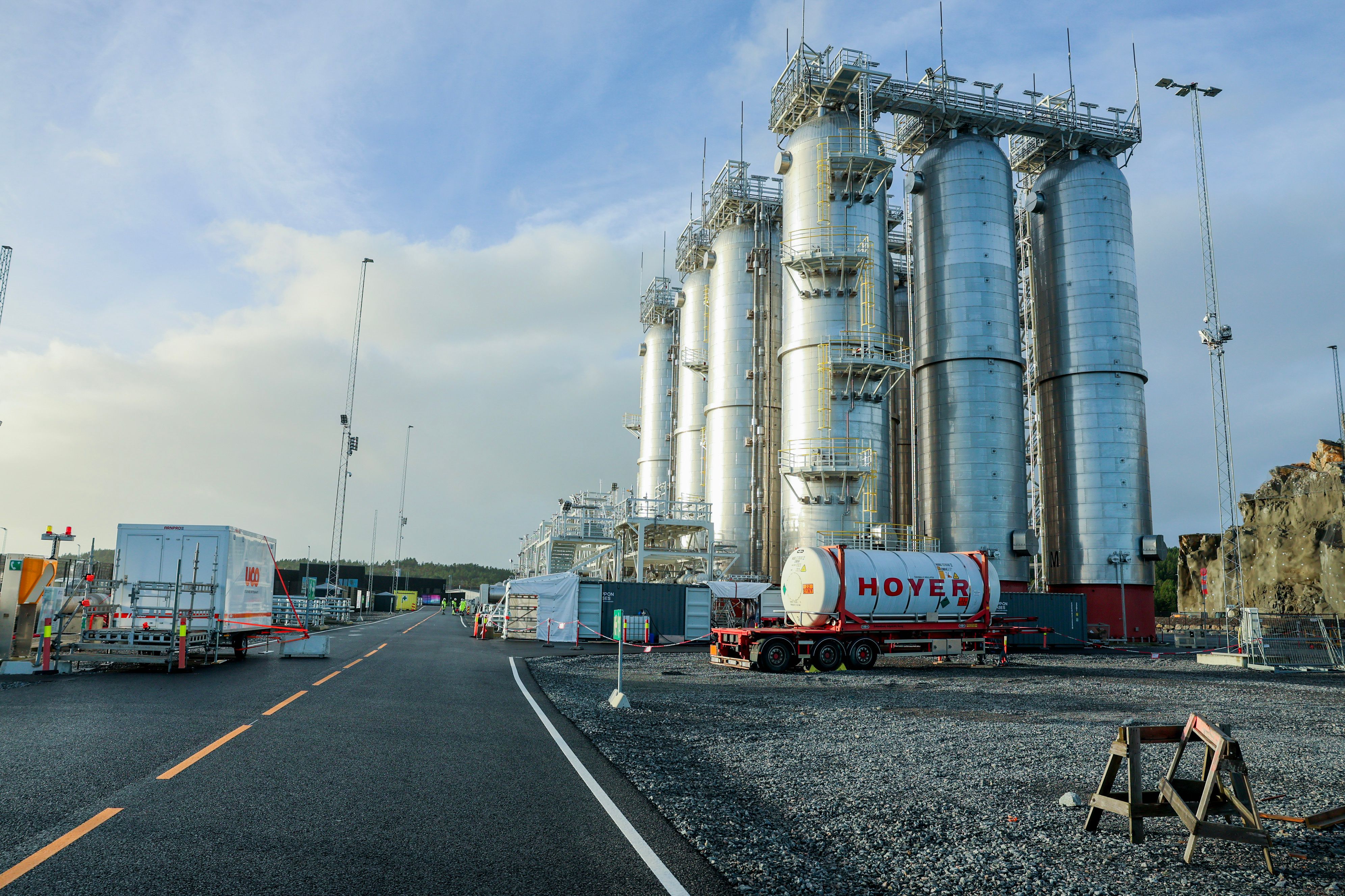Et industriområde med høye, sylindriske metalltanker, en vei og en hvit tankbil.