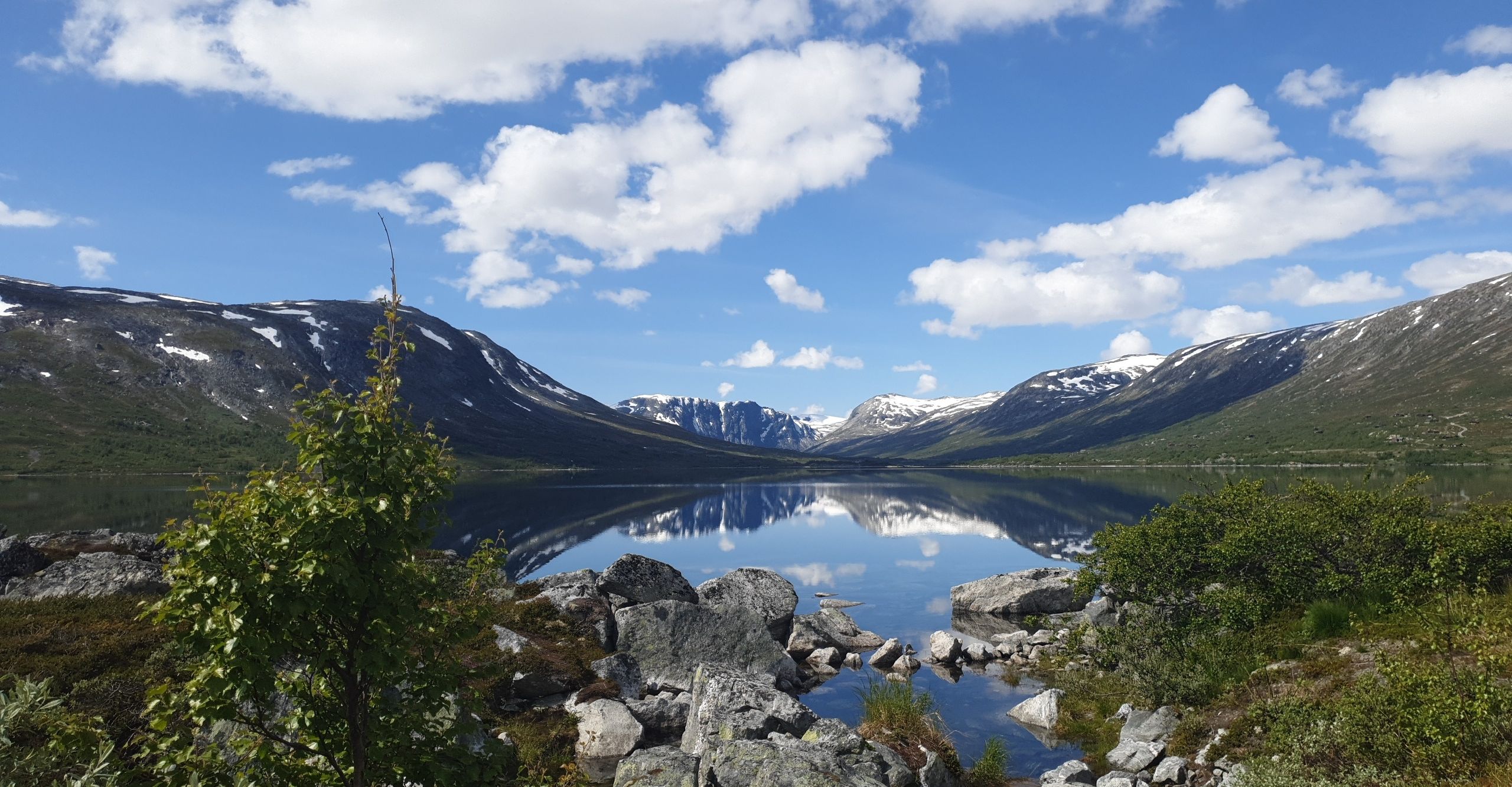 Rolig innsjø som speiler fjell med snøflekker og blå, skyet himmel, med steiner og sparsom vegetasjon i forgrunnen.