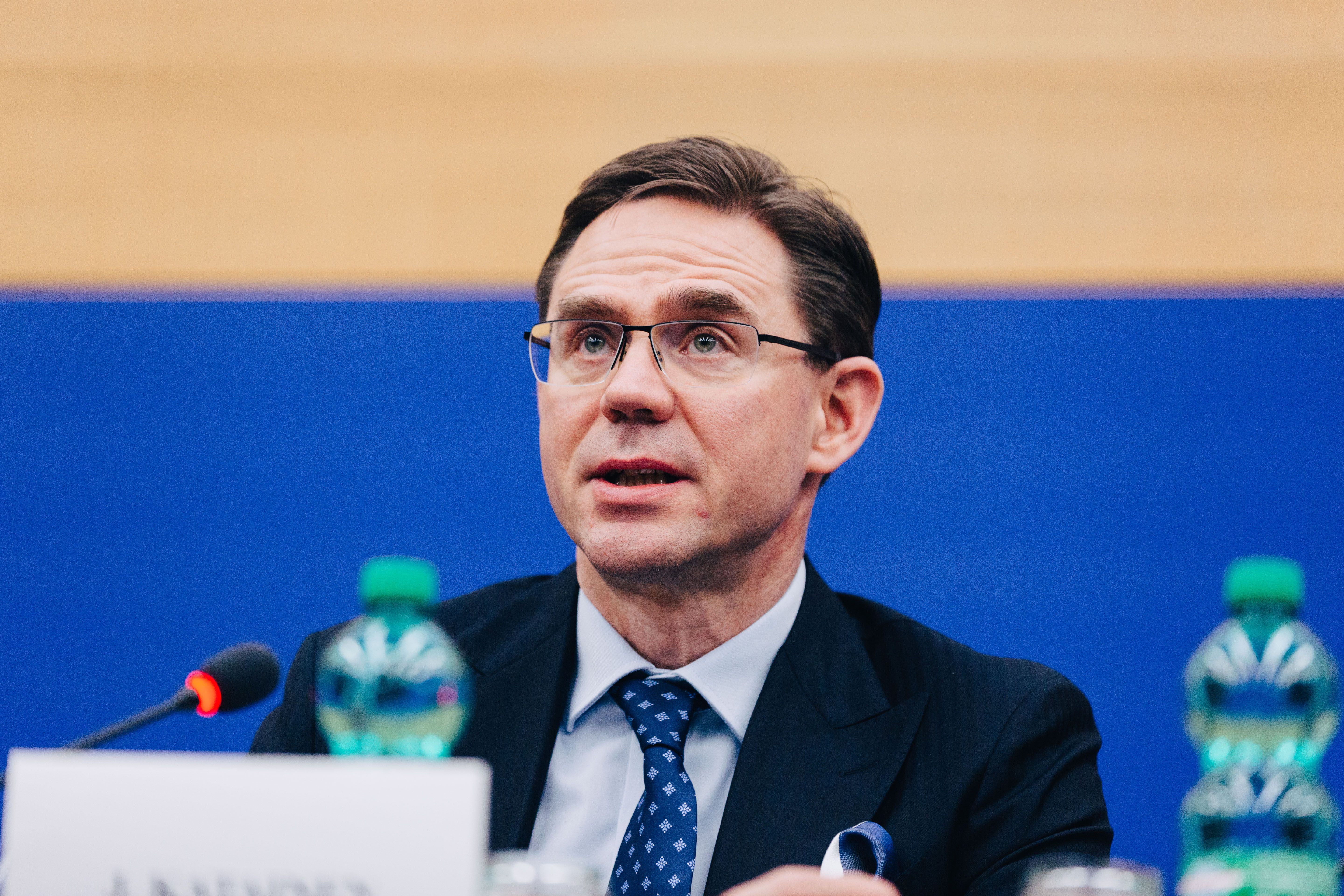 A man in a suit and glasses speaks into a microphone at a table with water bottles.