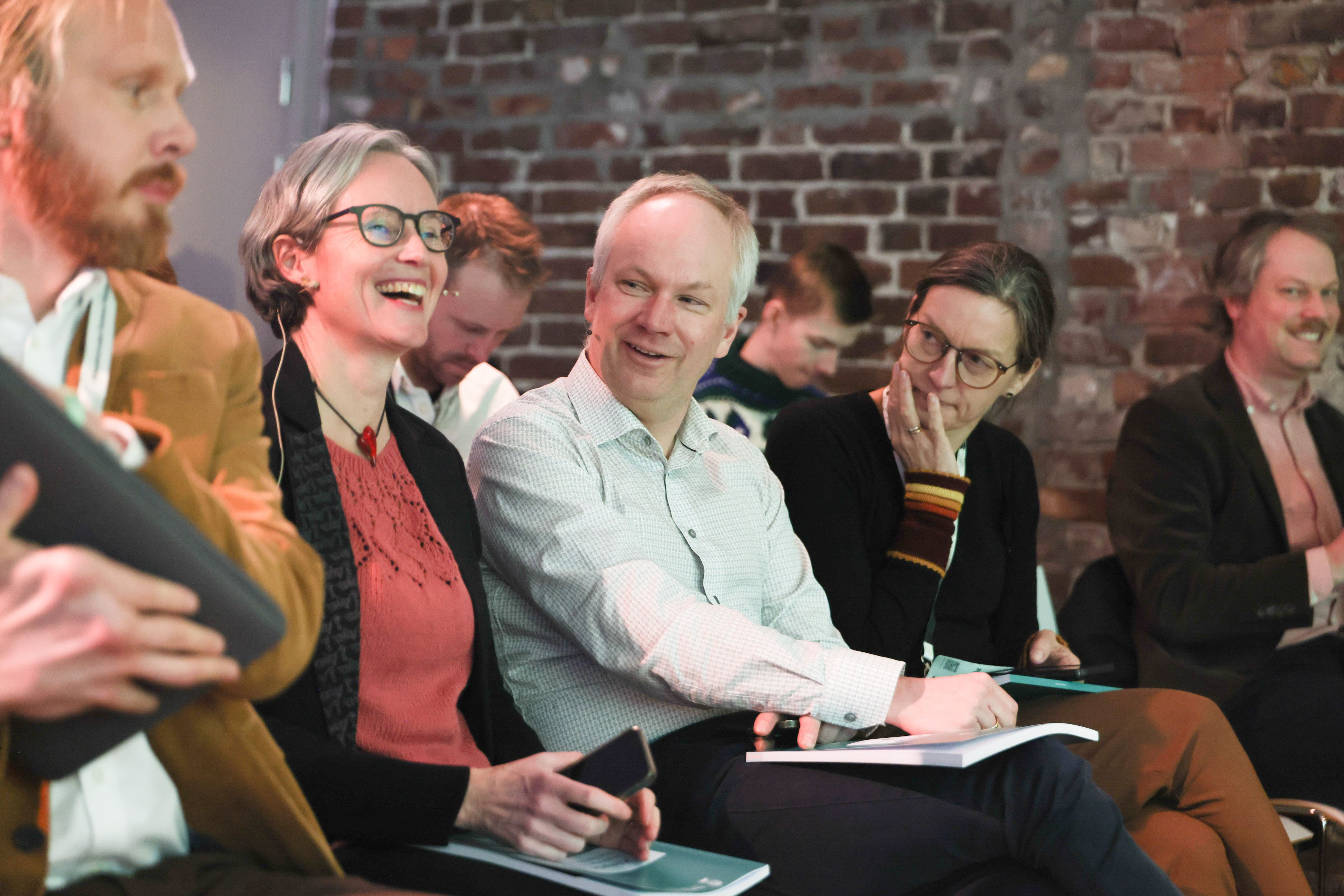 En gruppe ulike voksne som smiler og ler mens de sitter sammen i en uformell setting.