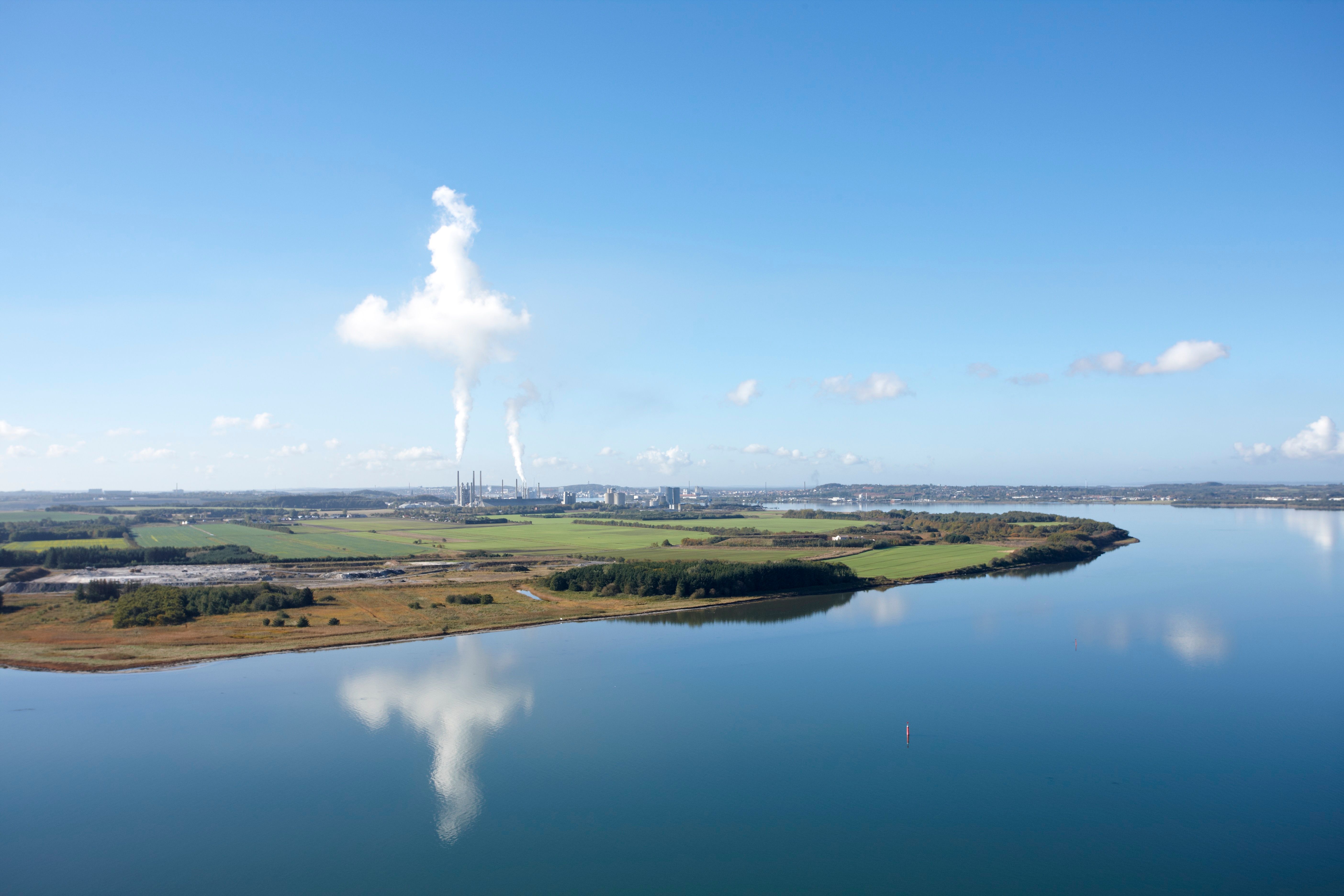 Et industrianlegg med rykende piper på en halvøy, speilet i rolig blått vann.