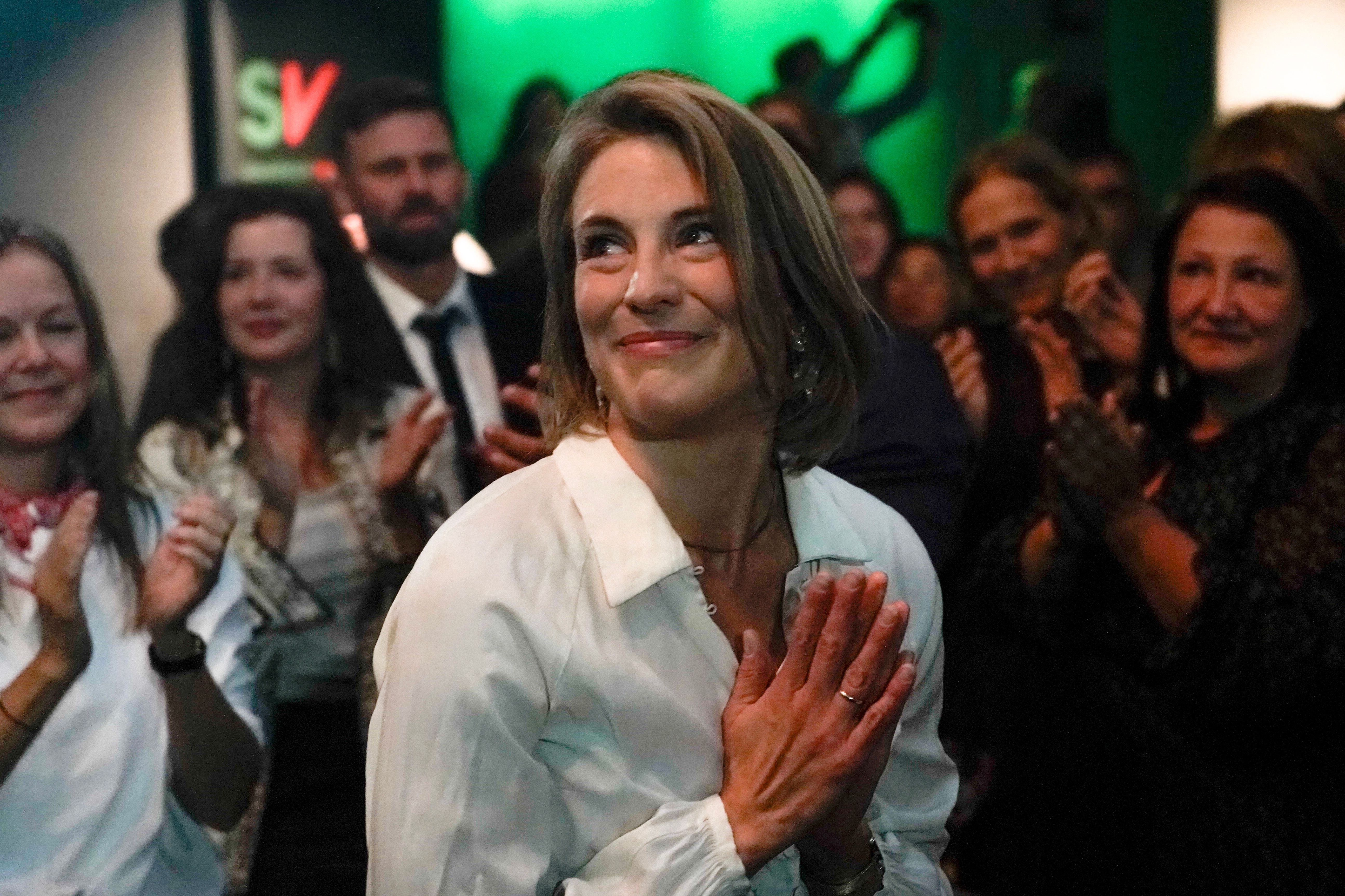 En smilende kvinne i hvit skjorte står med hendene foldet foran seg, foran en appluderende folkemengde og et grønt «SV»-skilt.