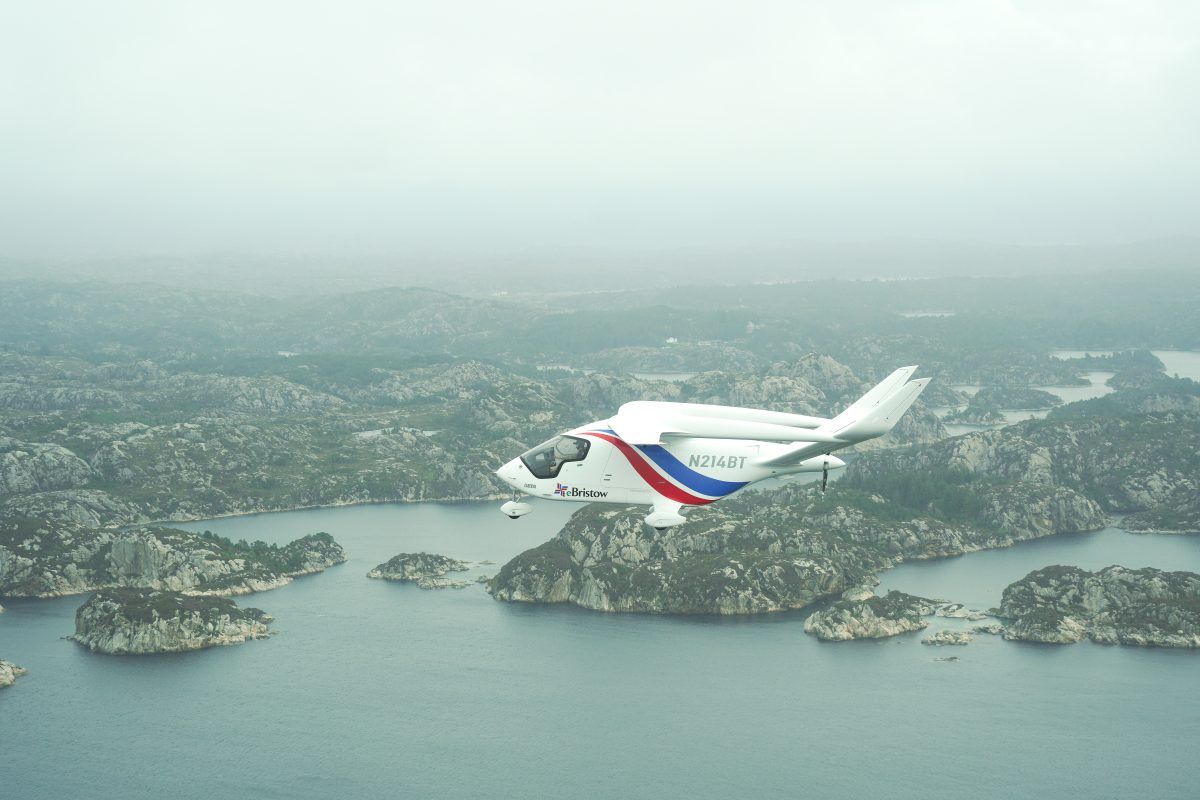 Et hvitt, elektrisk fly med røde og blå aksenter flyr over et tåkete landskap av steinete øyer og vann.