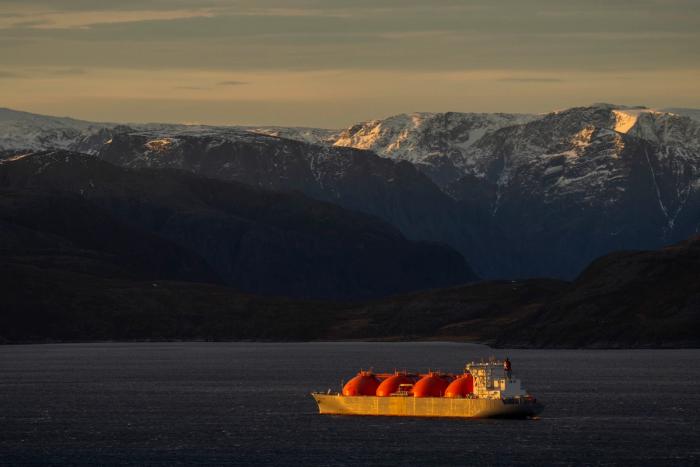 Et LNG-skip med glødende oransje tanker seiler på mørkt vann med snødekte fjell i bakgrunnen.