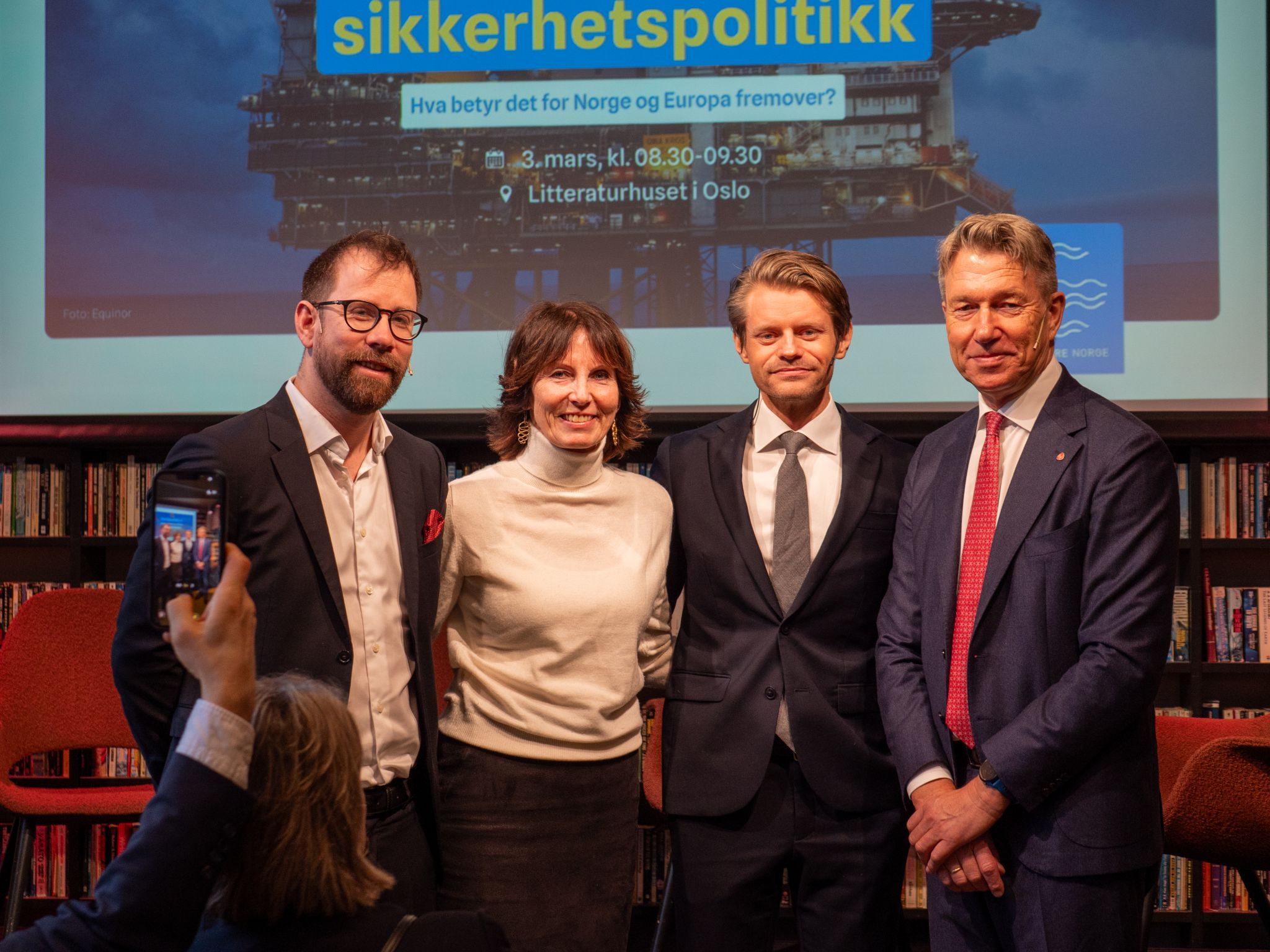 Fire personer, tre menn og én kvinne, poserer for et bilde på et arrangement med et bilde av en oljerigg og tekst om sikkerhetspolitikk på en stor skjerm bak dem.
