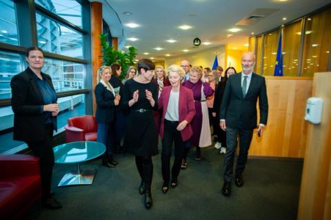 Ursula von der Leyen walks and smiles with a woman in black and a man in a suit, followed by others, in a modern building with EU flags.