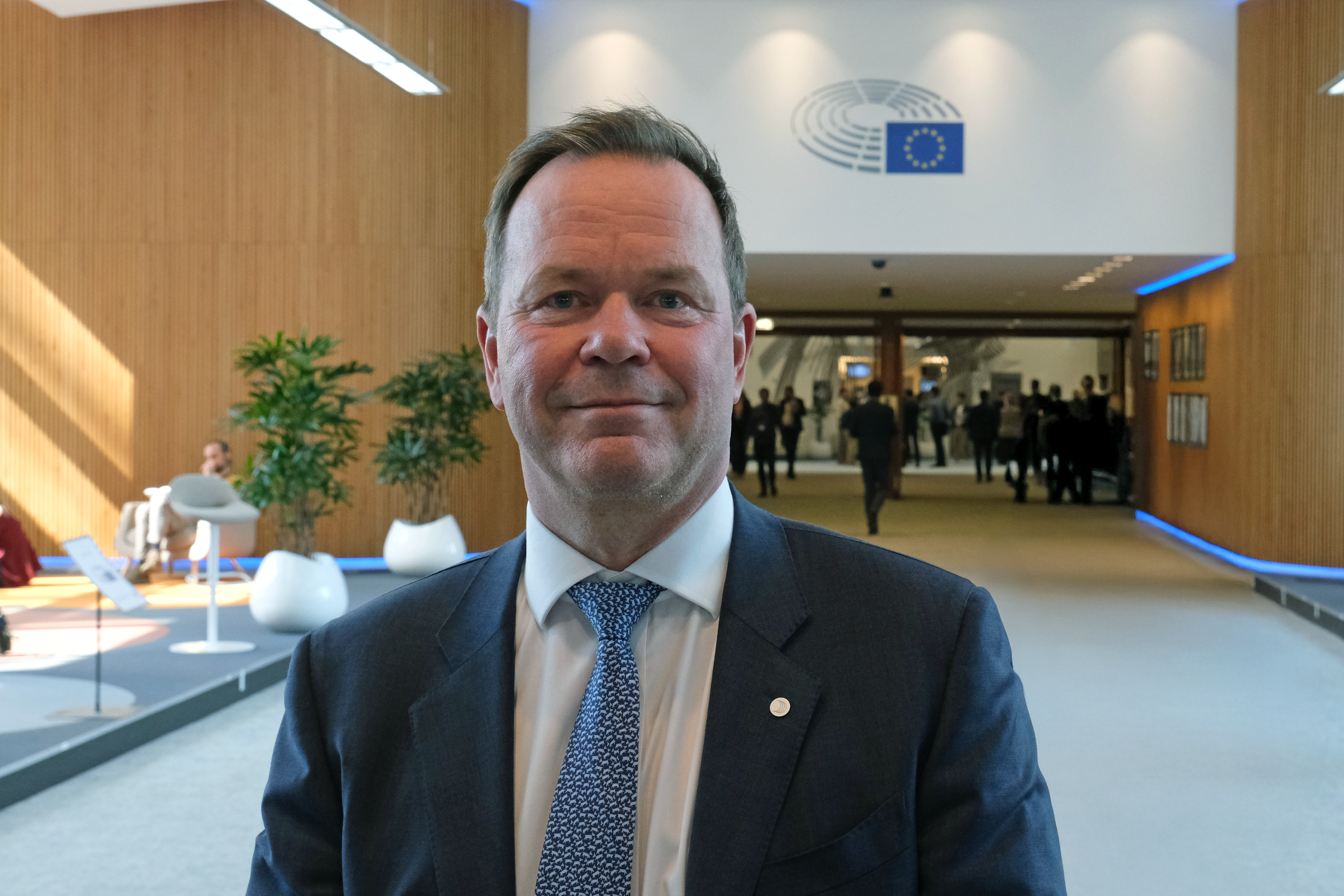 A man in a suit and tie smiles in a modern building, with the European Parliament logo visible in the background.