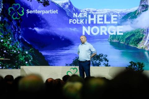 En mann står avslappet på en scene foran en skjerm som viser en norsk fjord, Senterpartiets logo og slagordet «NÆR FOLK I HELE NORGE».