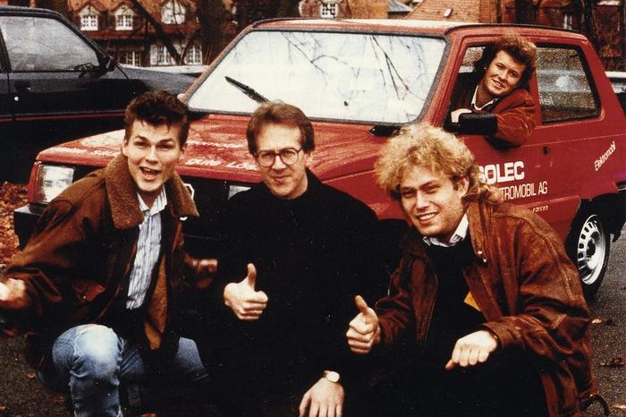 Fire smilende menn poserer med en rød SOLEC-elbil, to av dem viser tommel opp.