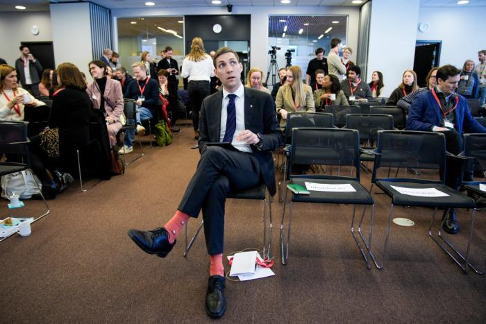 En mann i dress og knalloransje sokker sitter i et møterom og ser opp, med andre deltakere og mange tomme stoler rundt seg.